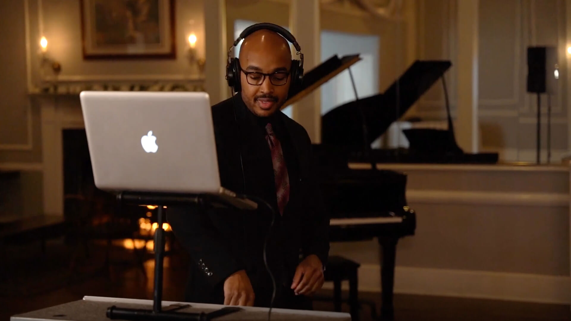 A man wearing headphones and a suit stands behind a laptop on a stand, with a grand piano and elegant décor visible in the background. He appears to be working with audio equipment.