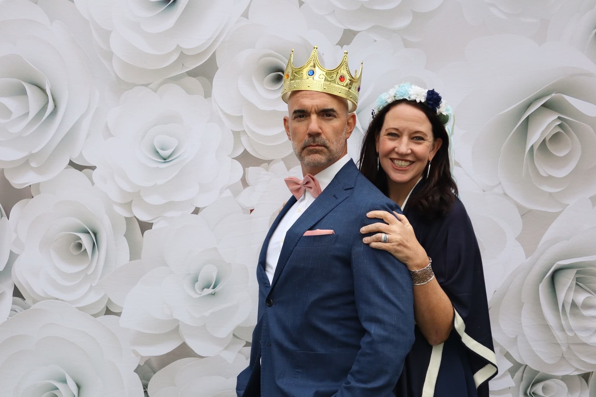 A man in a blue suit and gold crown stands in front of a white paper flower backdrop. A woman wearing a flower crown and dark shawl smiles and stands behind him with her hand on his shoulder.