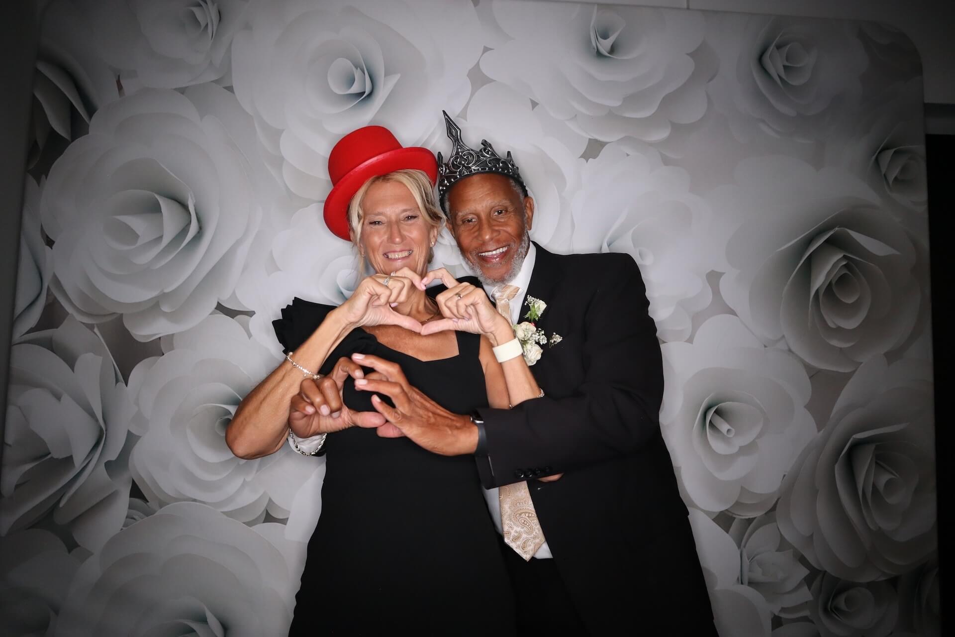 A woman in a red hat and a man in a silver crown pose in front of a white rose backdrop, both smiling and forming a heart shape with their hands. The man is wearing a suit and boutonniere.