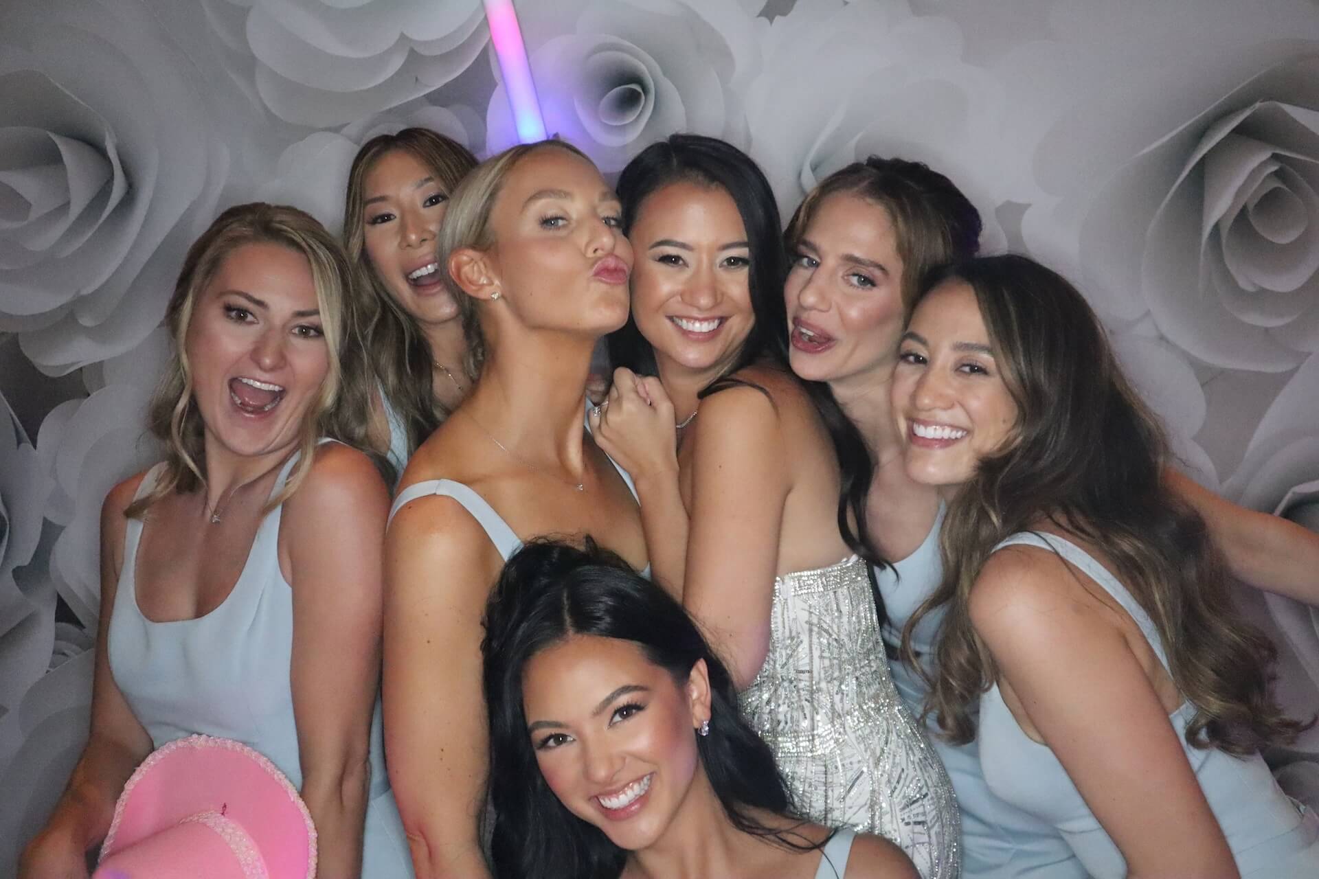 Seven women pose closely together in front of a floral backdrop, most wearing light blue dresses. One wears a headband with a light-up unicorn horn. They are smiling and appear to be at a celebratory event.