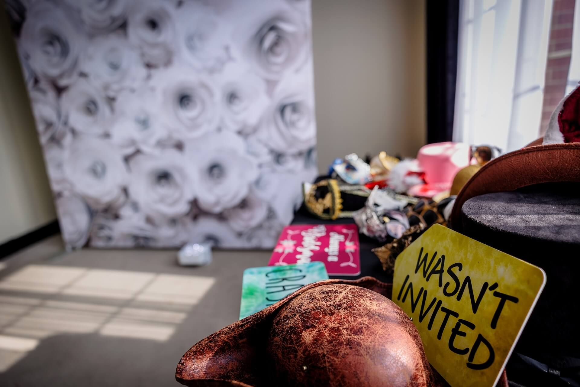 A table with assorted hats and props sits near a window. In the foreground is a yellow sign reading “WASN’T INVITED.” A floral backdrop stands in the background with bright sunlight streaming in.