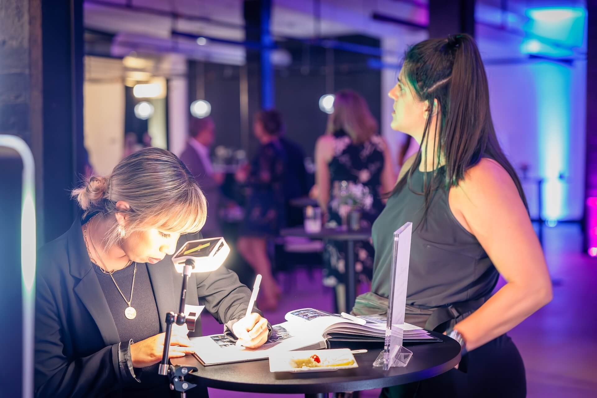 A woman sits at a small round table signing a book under a bright light, while another woman stands nearby watching. The background shows people socializing in a dimly lit room with colorful lighting.
