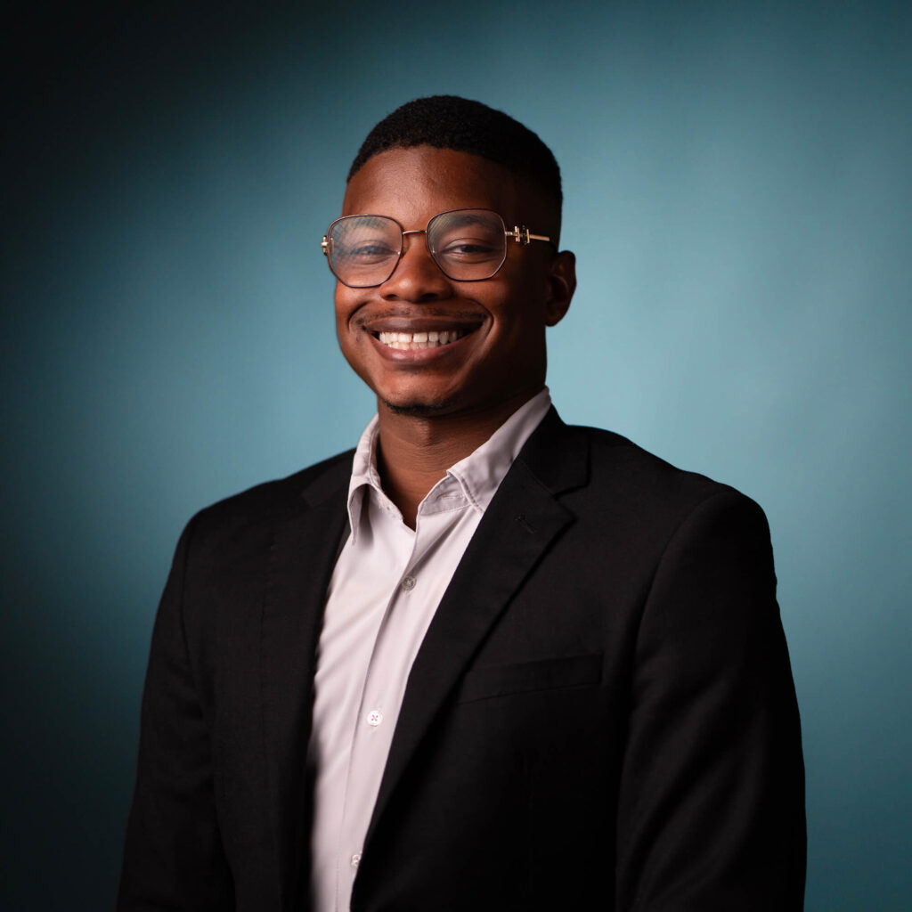 A person wearing glasses, a white shirt, and a black blazer is smiling in front of a solid teal background. The person is facing the camera in a professional portrait style.
