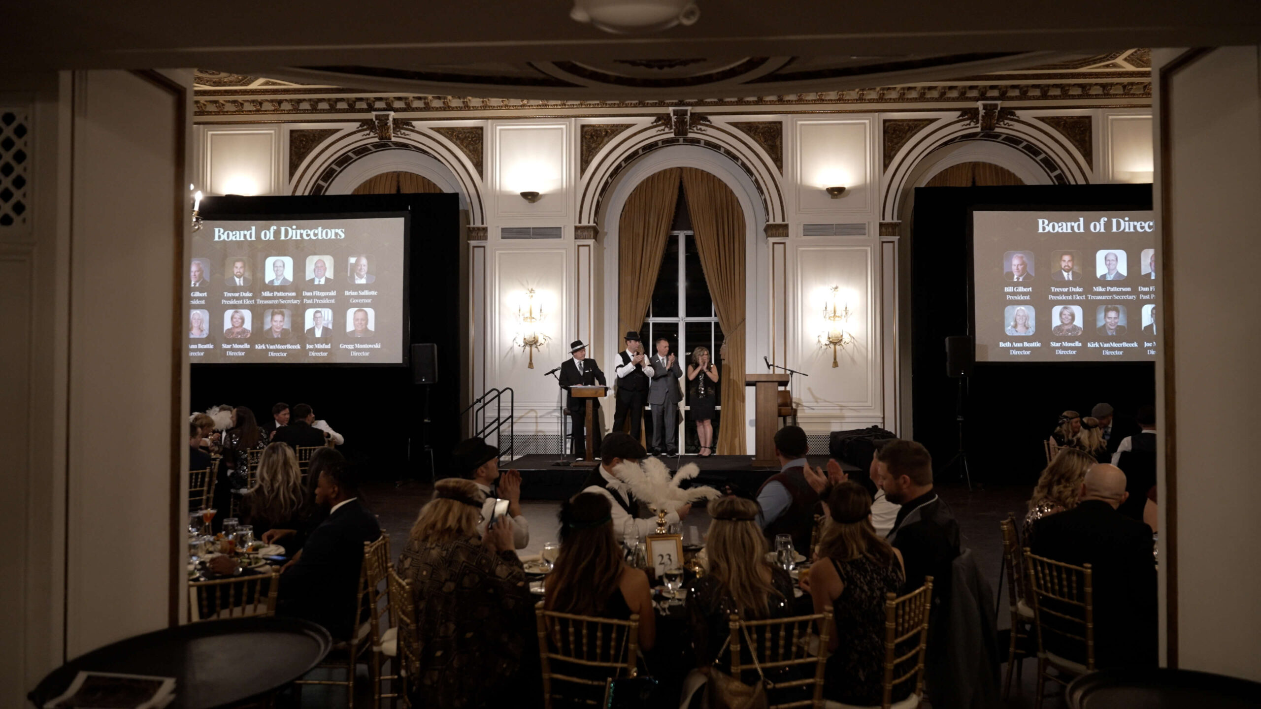 A formal banquet is taking place in an ornate hall. Attendees sit at round tables while three people stand on stage. Two screens display "Board of Directors" with photos and names. The room has arched windows and gold decor.