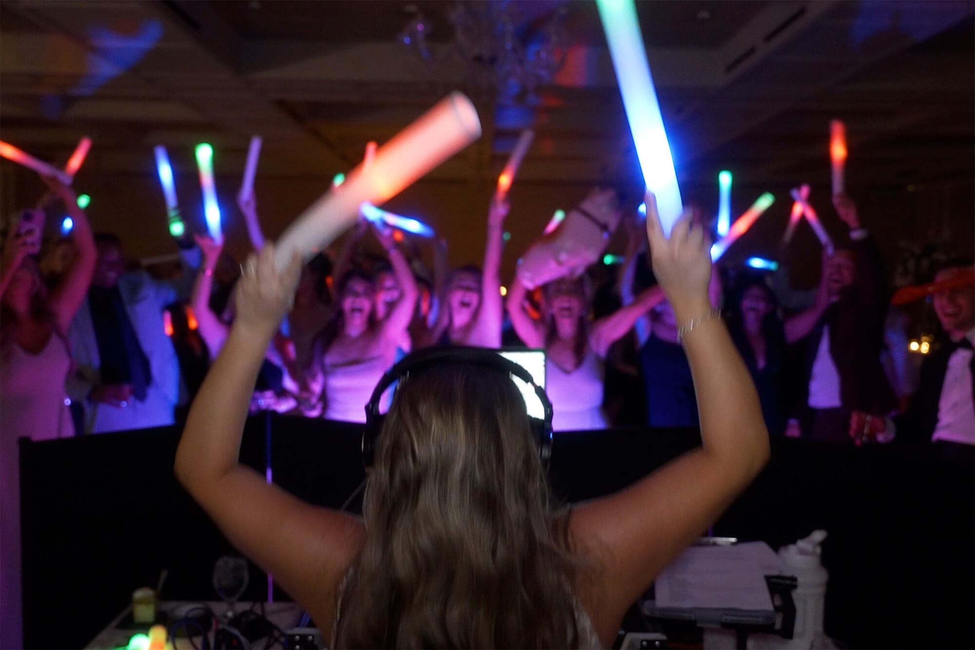 A DJ with long hair, seen from behind, holds up two light sticks while a crowd in a dimly lit room waves glowing sticks and dances, creating a colorful party atmosphere.