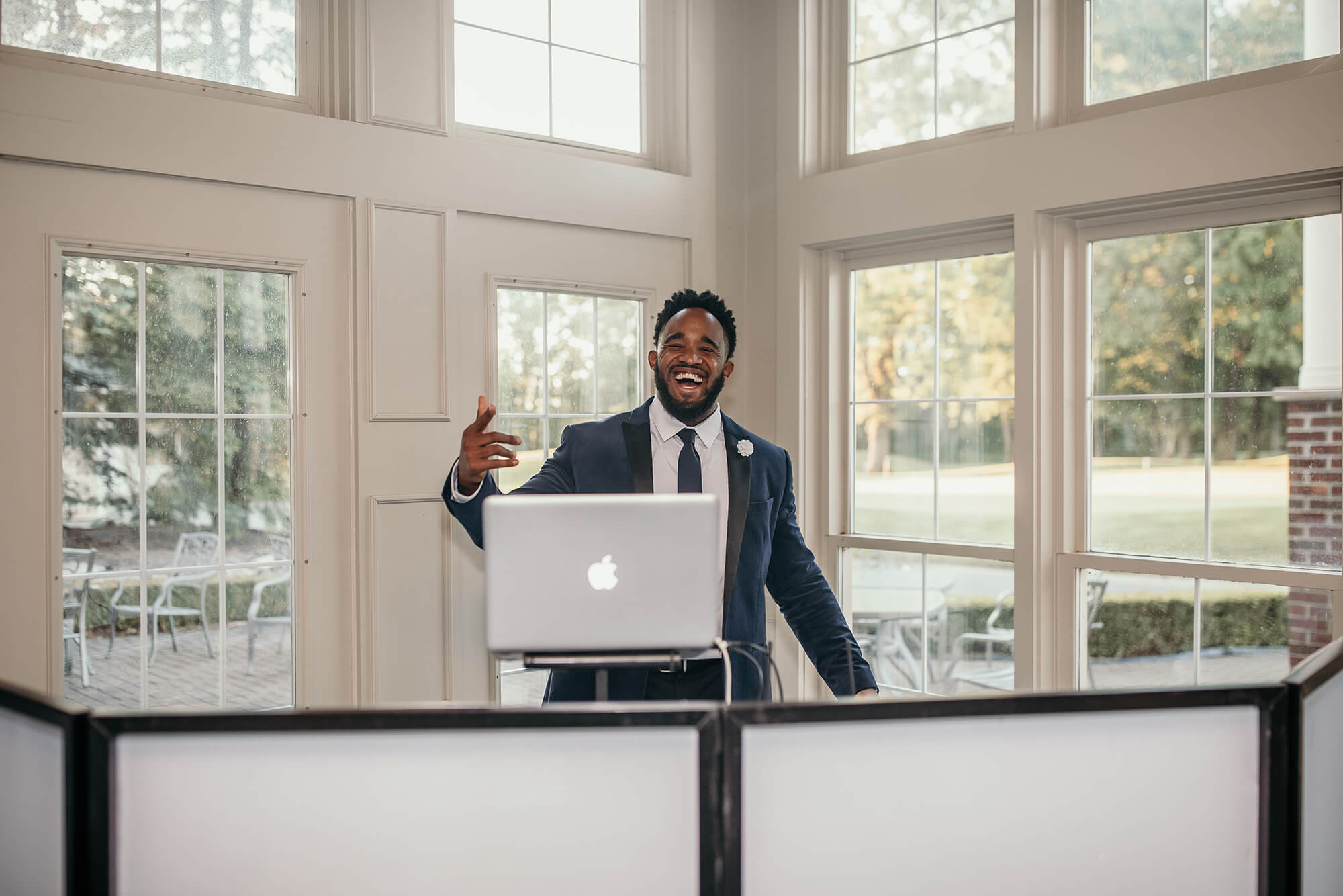 A man in a suit stands behind a laptop and monitors in a bright room with large windows, smiling and gesturing with his hand. Trees and outdoor furniture are visible outside.