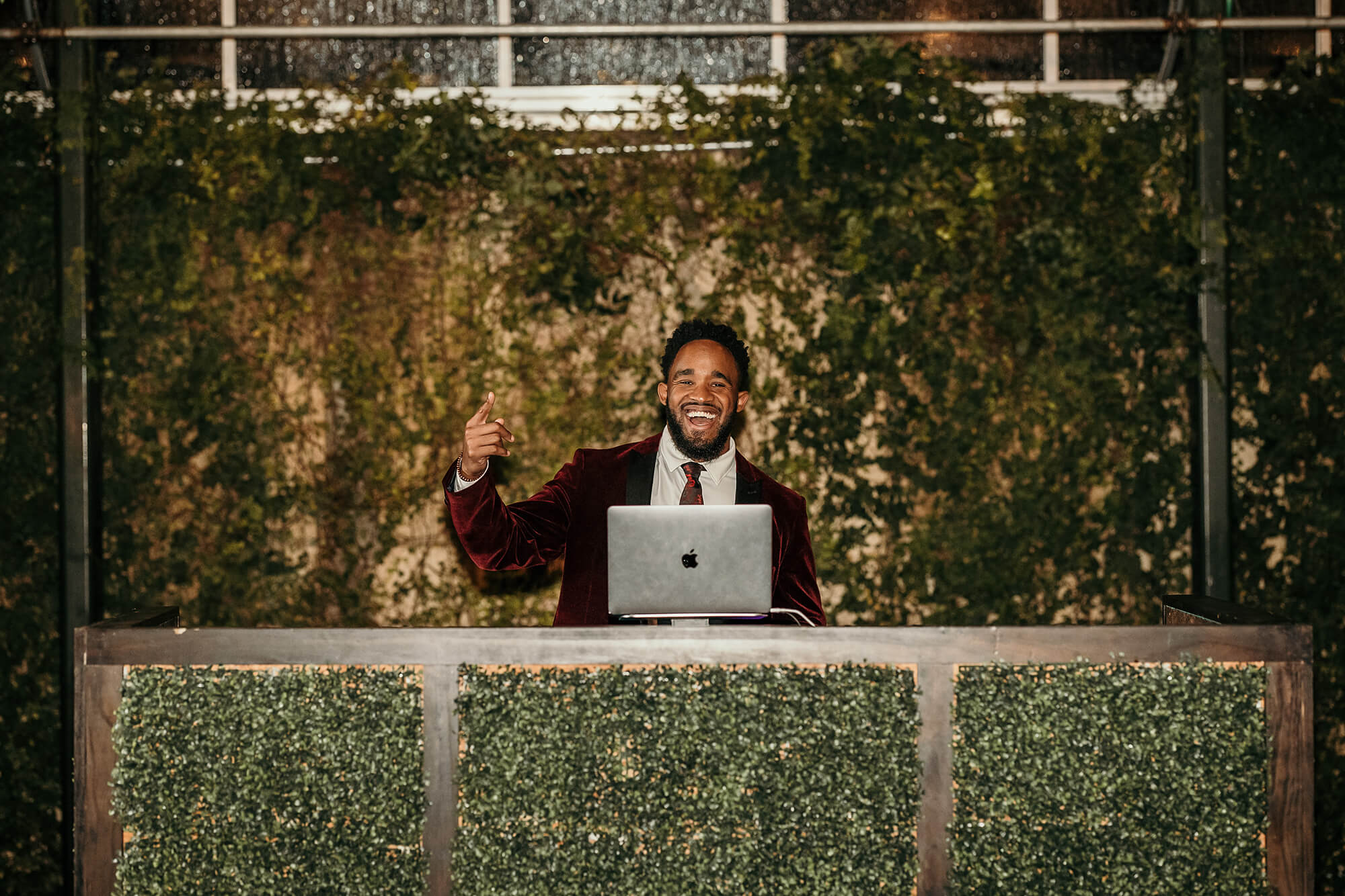 A man in a burgundy suit stands behind a modern DJ booth with artificial greenery panels, smiling and gesturing with one hand while using a laptop. The background is covered with leafy vines.