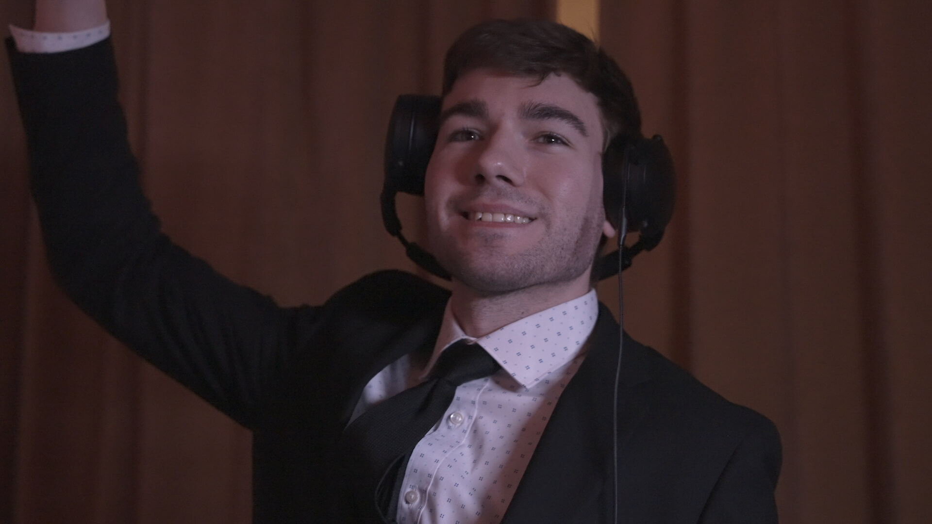 A man in a suit and tie smiles while wearing large headphones. He is raising one arm and standing in front of brown curtains.