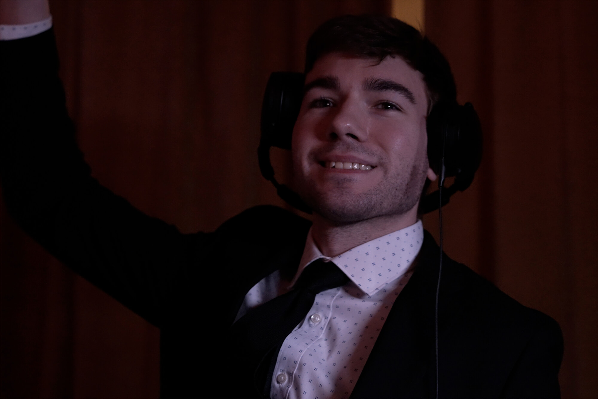A person in a suit and tie smiles at the camera while wearing large over-ear headphones, raising one arm. The background is dimly lit with a warm tone.