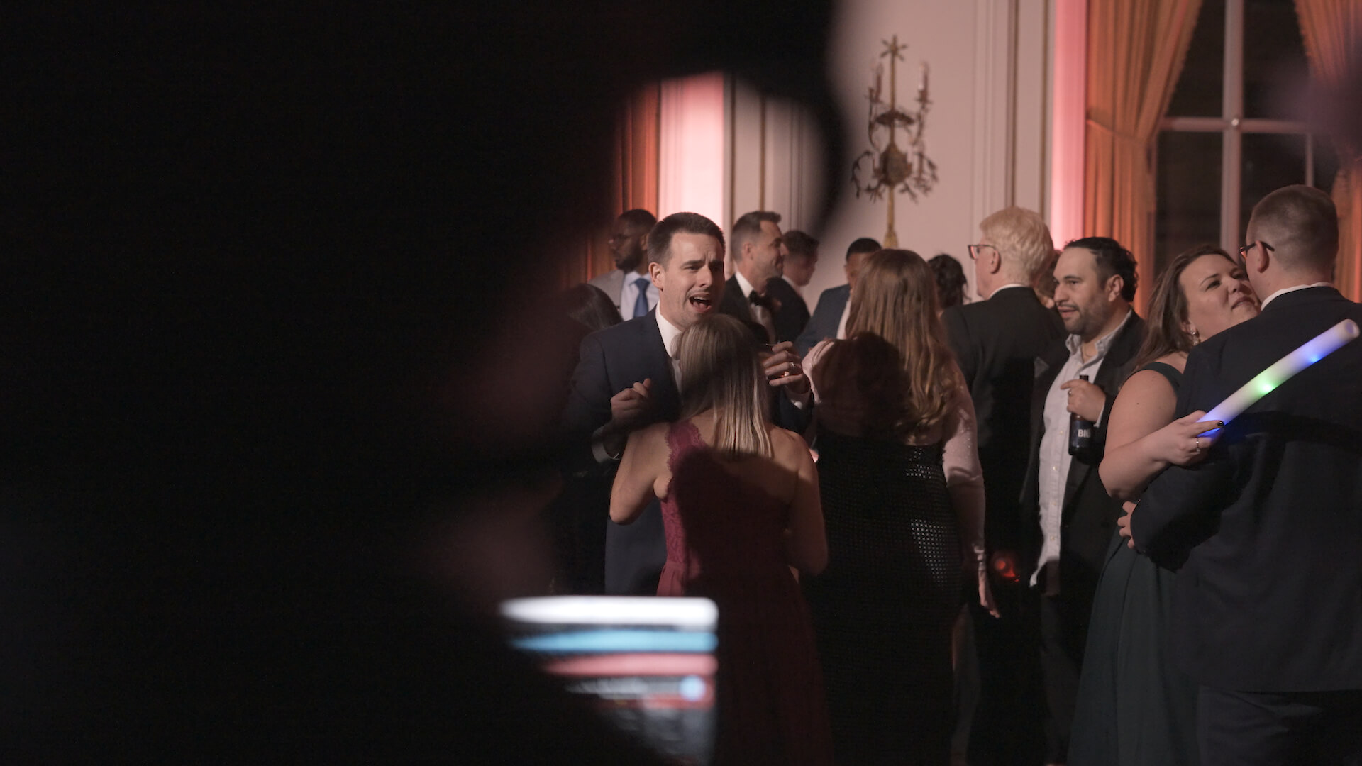 A group of people in formal attire socialize at an indoor event. Some hold drinks and talk in small groups. The foreground is partially obscured by a dark, out-of-focus shape. Warm lighting fills the room.