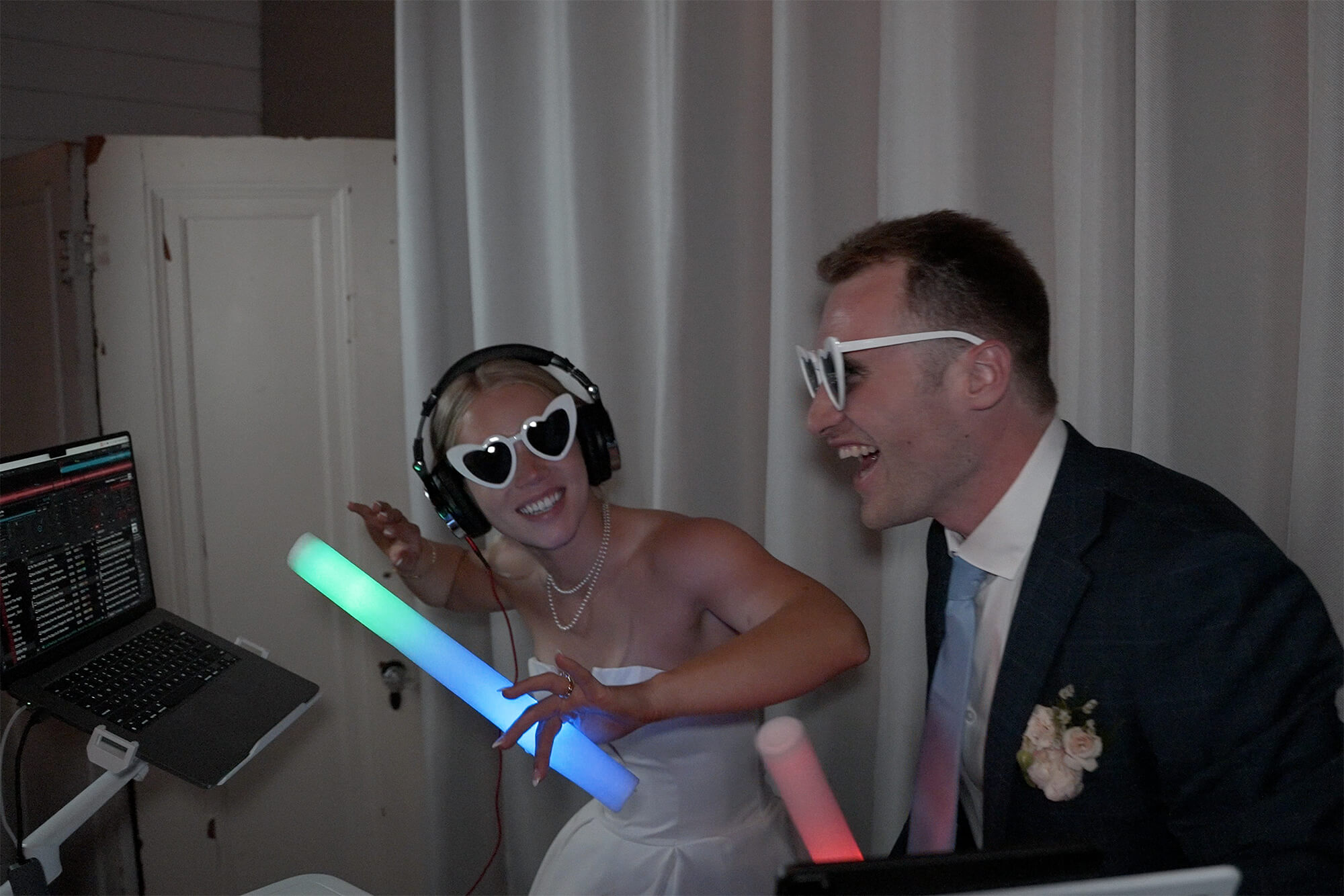 A woman in a white dress and a man in a suit wearing heart-shaped and oversized glasses are smiling and holding light-up sticks, standing next to DJ equipment with headphones on.