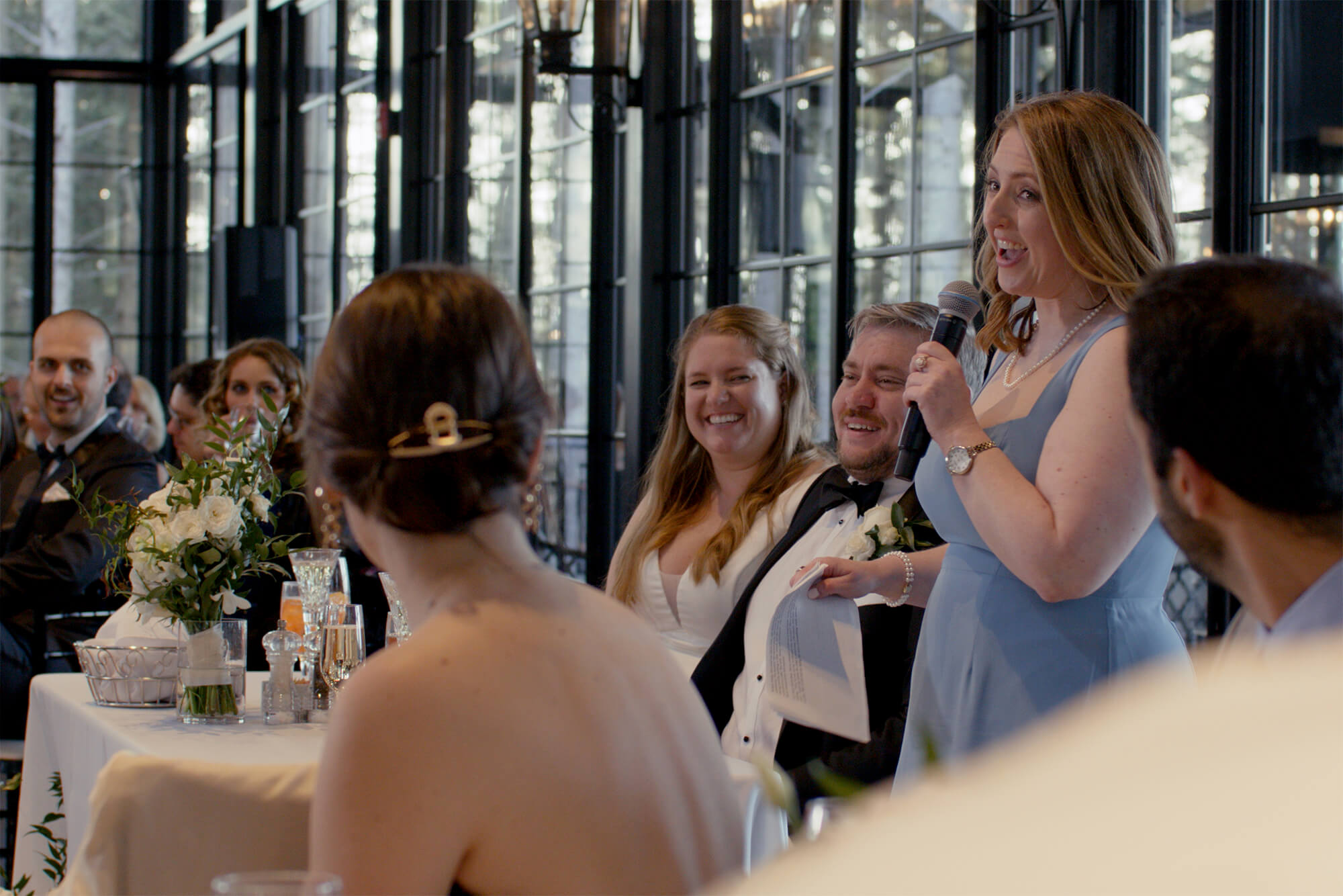A woman in a light blue dress stands and speaks into a microphone at a wedding reception, while guests and the bride and groom, who are seated at a table, smile and listen.