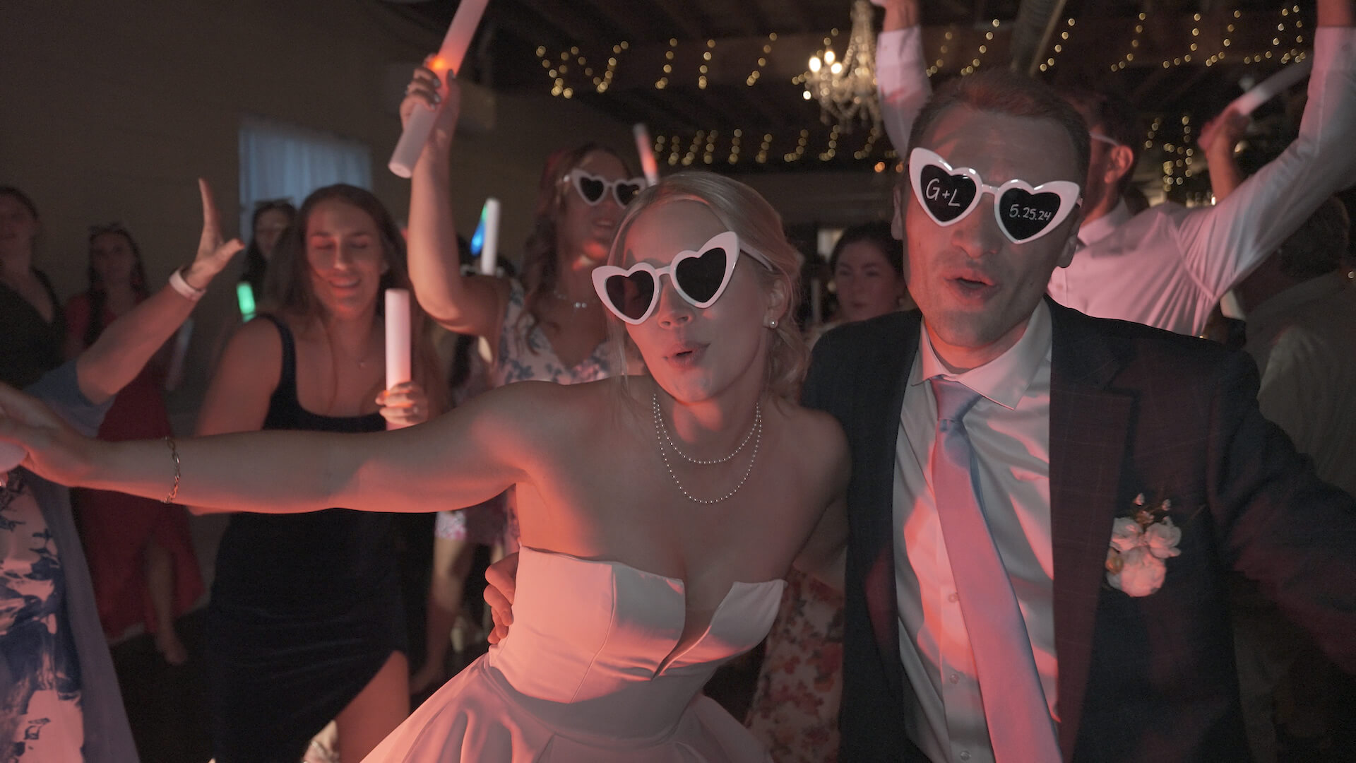 A bride and groom wearing heart-shaped sunglasses pose together on a dance floor, surrounded by guests holding glow sticks at a lively indoor wedding reception.