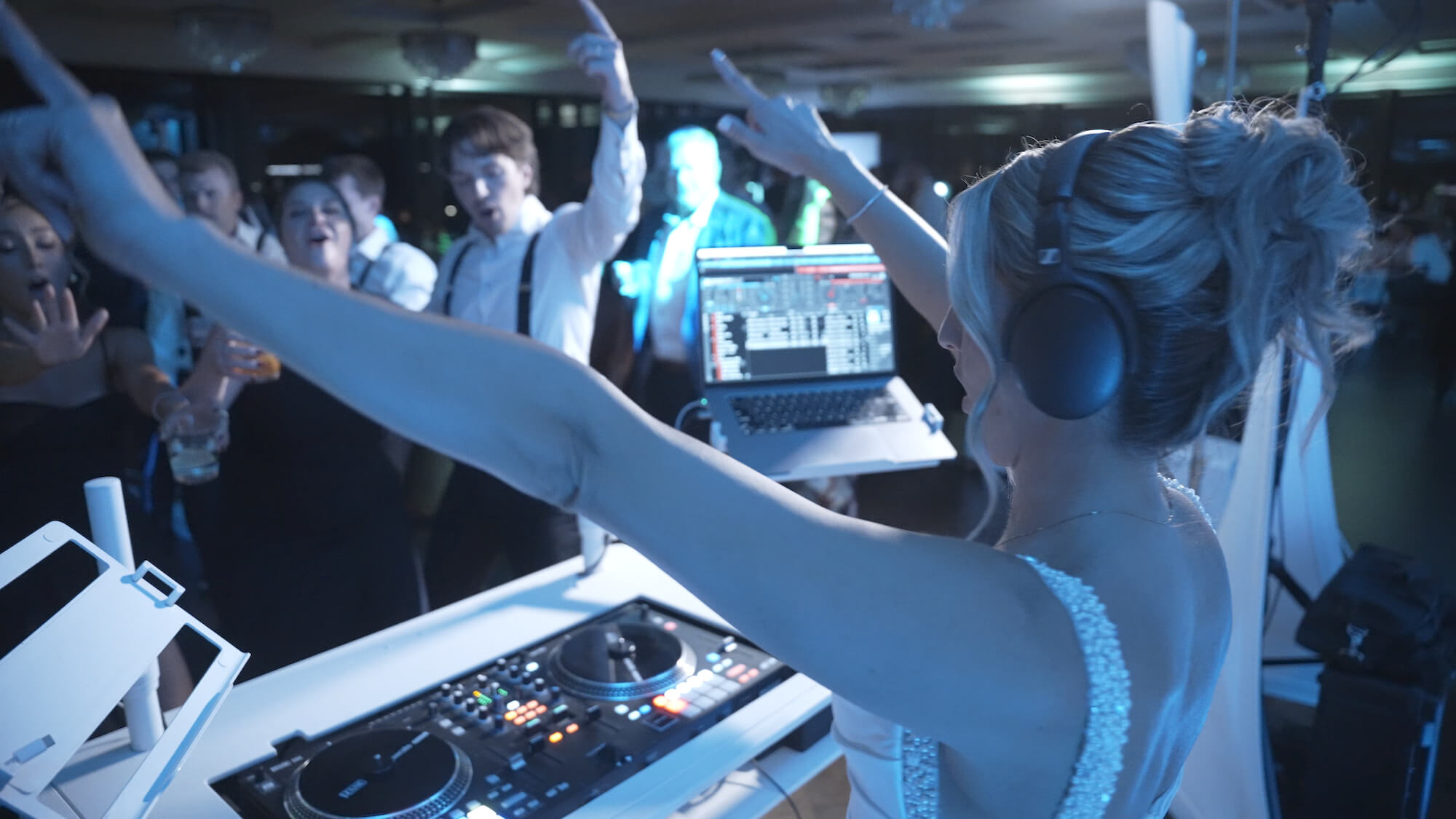 A female DJ with headphones stands at a DJ booth, raising her arms while guests on a crowded dance floor cheer and dance under blue lighting. A laptop and DJ controller are visible on the table.