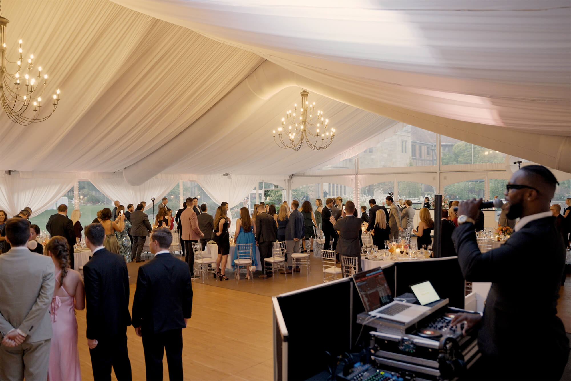 A large group of people stands inside a white tent with chandeliers, gathered around tables and a dance floor. A DJ with equipment is in the foreground, and natural light comes through the tent windows.