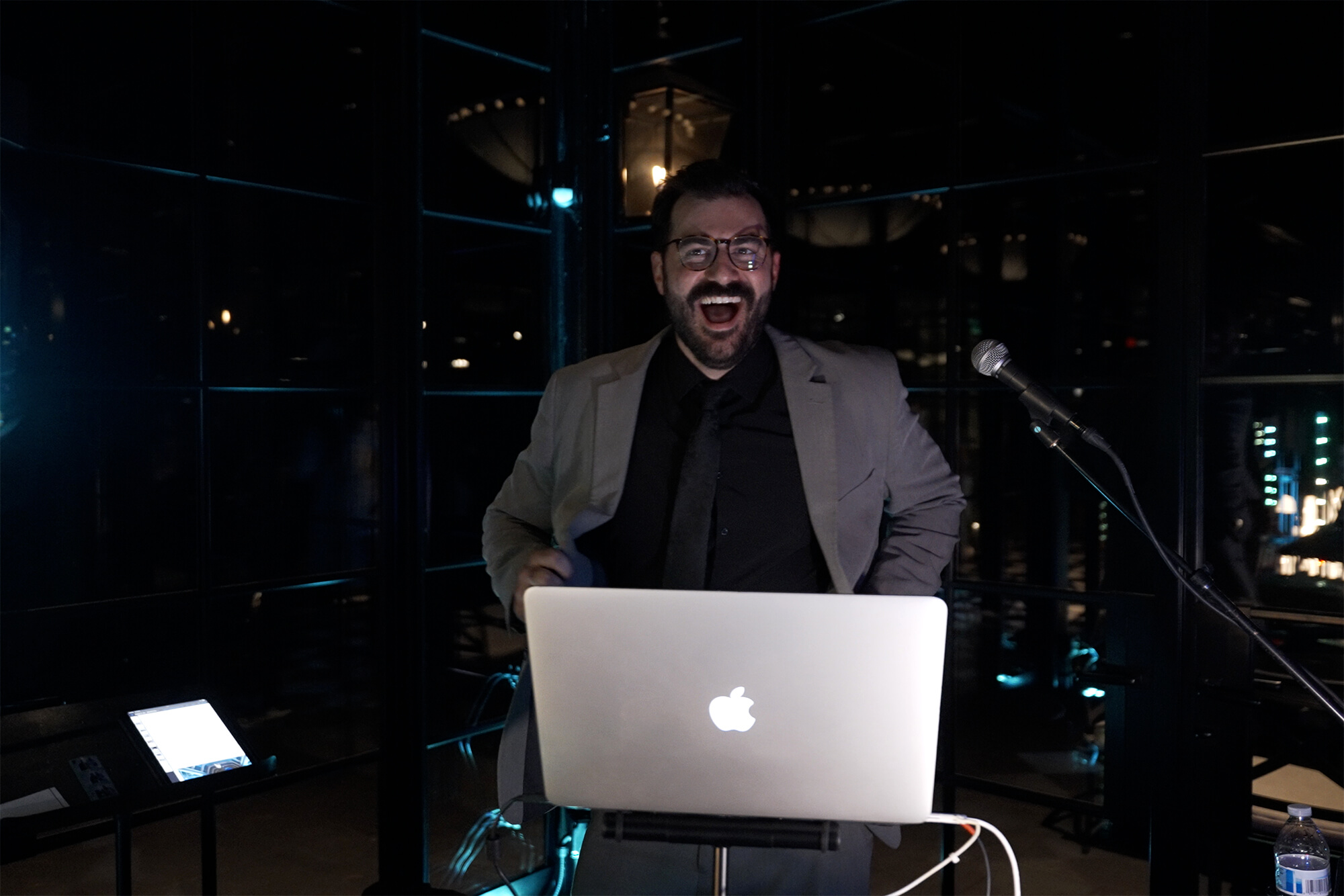 A man in a gray suit and black shirt stands behind a laptop with an Apple logo, smiling widely. He is beside a microphone in a dark room with reflections and blue accent lighting.