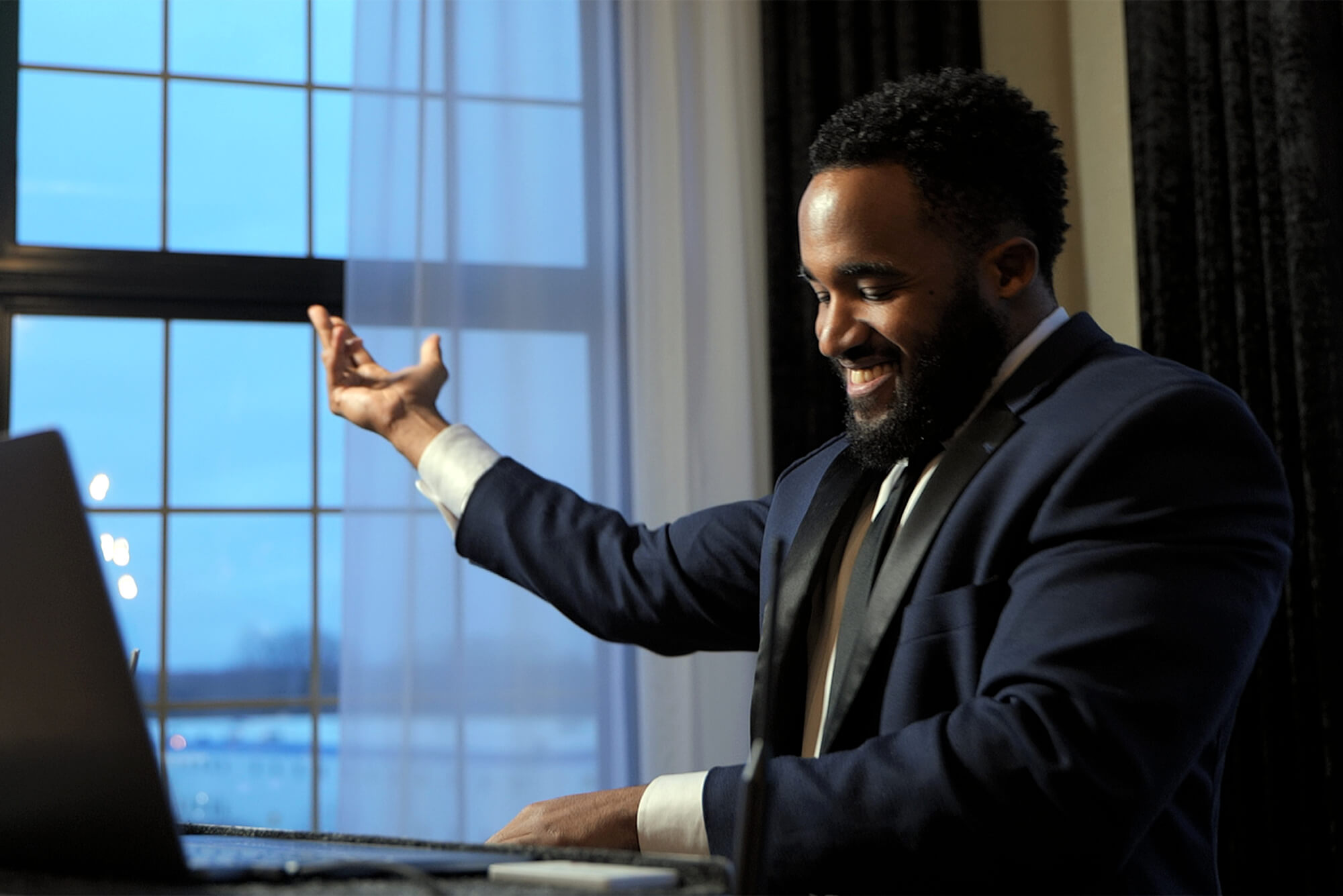 A man in a suit sits at a desk in front of a laptop, smiling and gesturing with one hand raised. A large window with curtains is in the background.