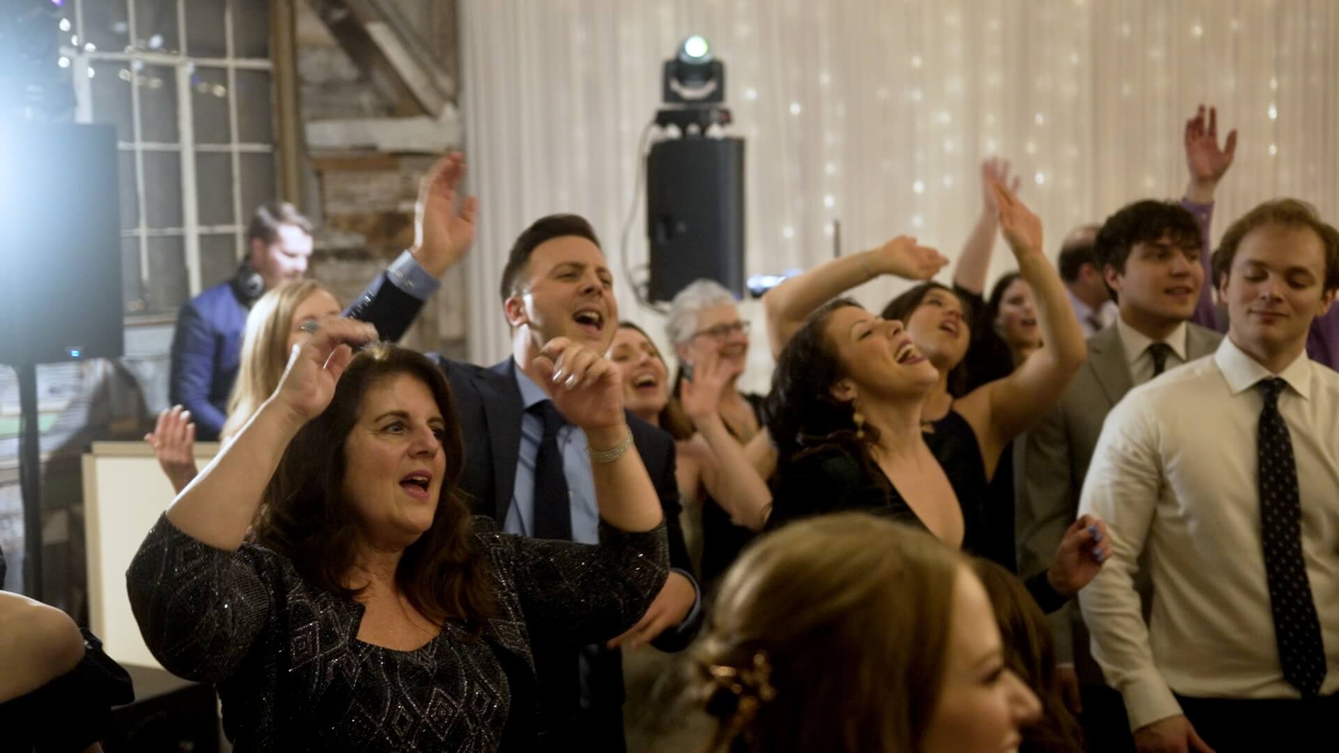 A group of people dressed in semi-formal attire dance and sing with their hands raised at an indoor event, with string lights and a DJ in the background.