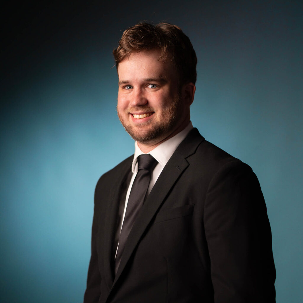 A man with light brown hair and a beard is wearing a black suit, white shirt, and black tie. He is smiling and posing in front of a plain blue background.