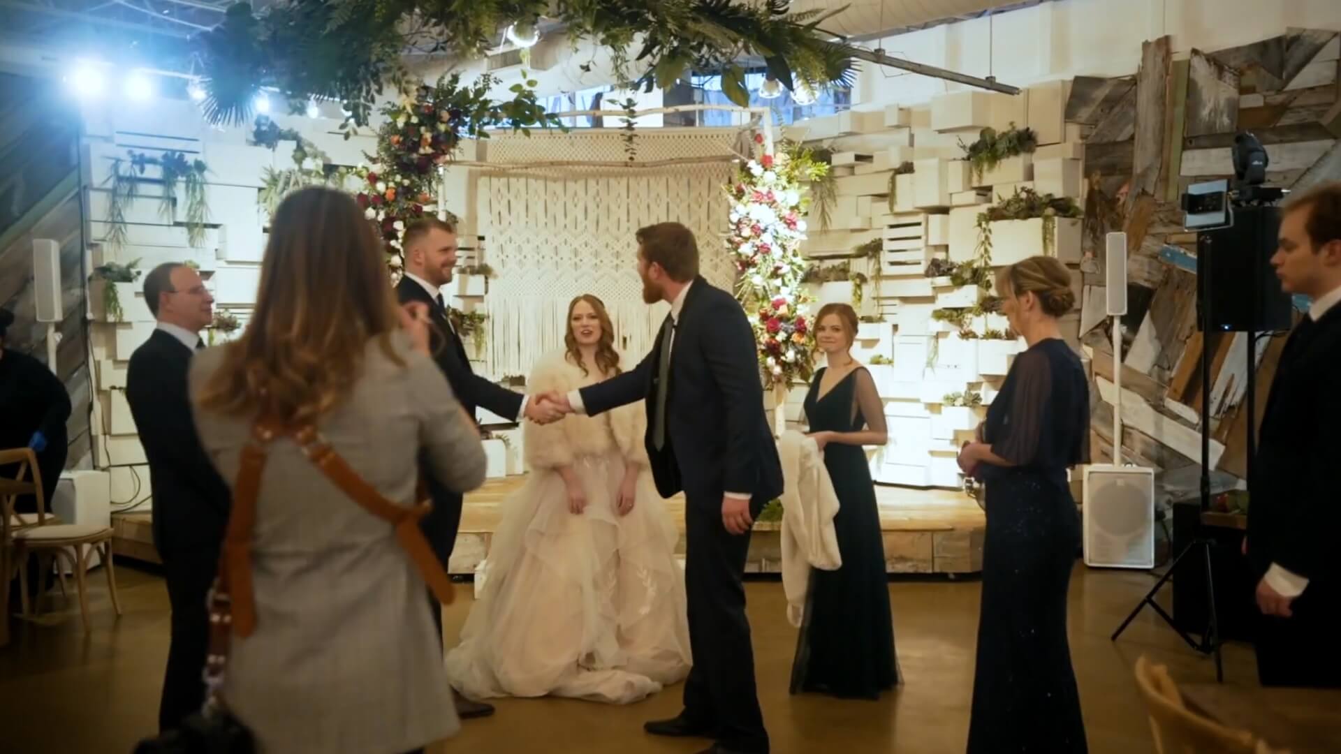 A bride in a white dress sits on a bench, smiling, while two men in suits shake hands in front of her. Several other people in formal attire stand around them in a decorated indoor venue.