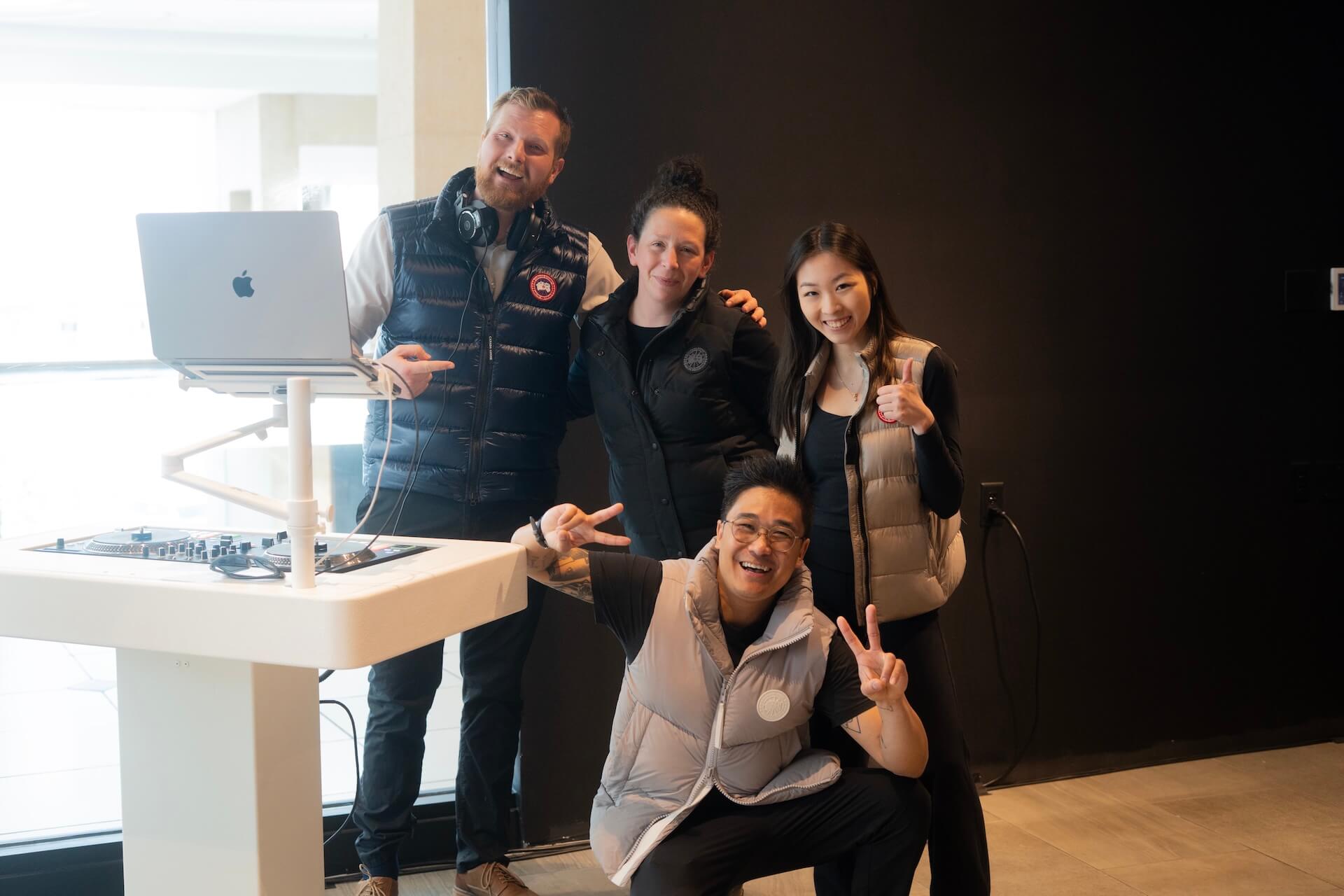 Four people pose and smile in front of a black wall and white DJ table with a laptop. Three stand while one kneels in front, holding up peace signs. All wear dark pants and light-colored vests.