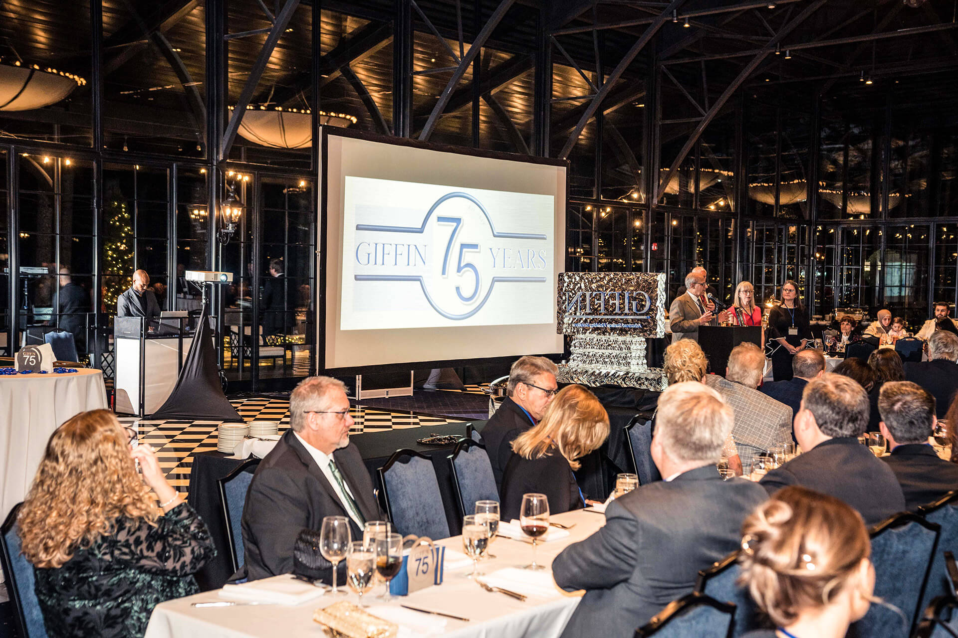 A formal event is taking place in a large, glass-walled venue. Attendees are seated at tables facing a screen displaying "Giffin 75 Years." Several people are speaking at a podium with a decorative ice sculpture nearby.