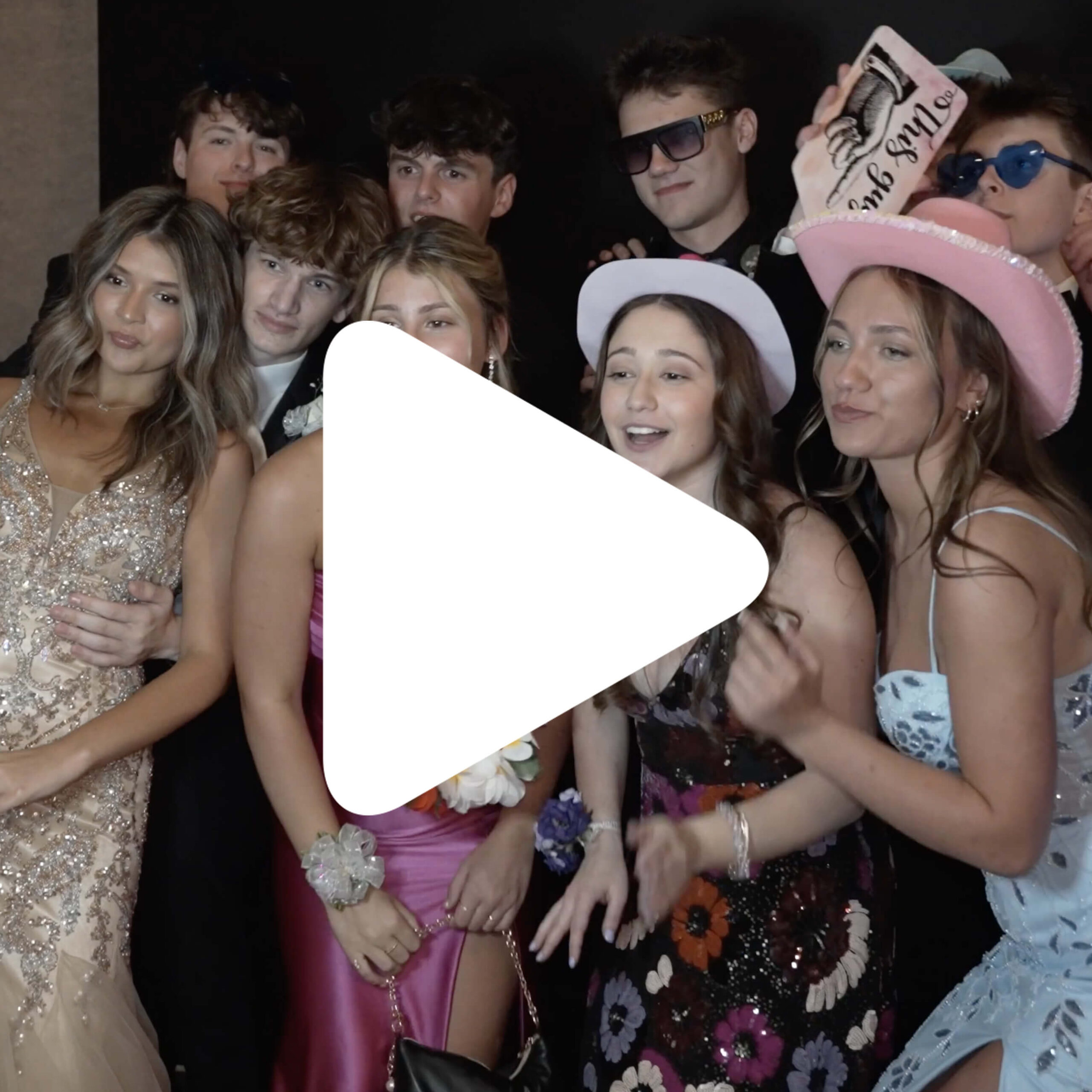 A group of young people dressed in formal and semi-formal outfits pose together for a photo; some are wearing hats and sunglasses. A large white play button is centered over the image.