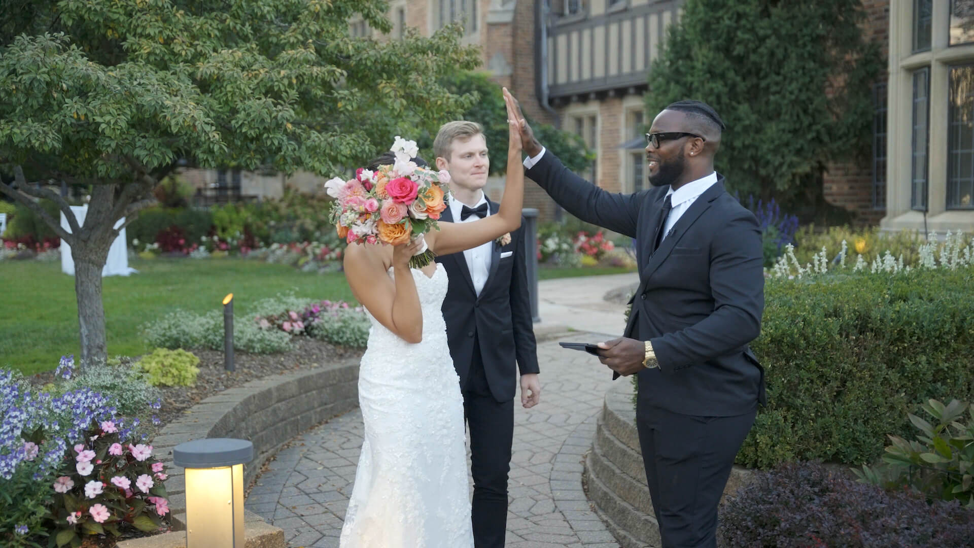 A bride in a white wedding dress covers her face with a bouquet while a groom in a tuxedo stands beside her. A man in a black suit gives the groom a high five outside in a garden setting.