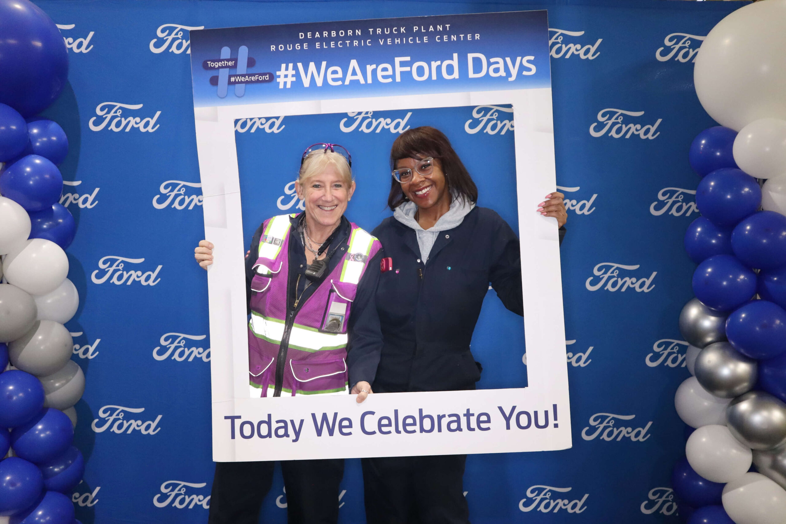 Two people stand smiling inside a large photo frame with the words "#WeAreFordDays" and "Today We Celebrate You!" on it, in front of a blue Ford backdrop with balloon decorations on both sides.