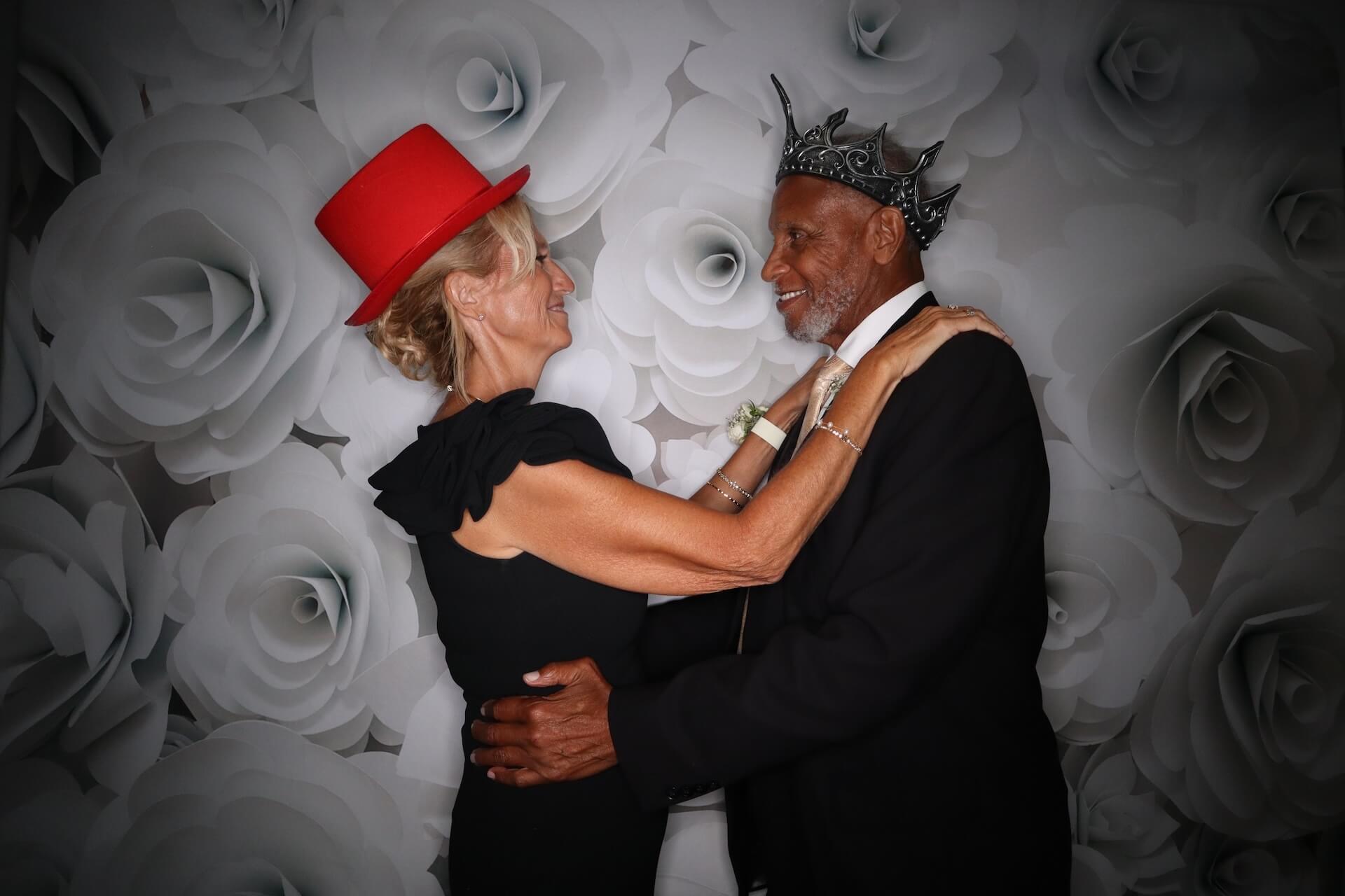 A woman in a red hat and a man in a silver crown dance together, smiling at each other, in front of a backdrop printed with large white roses.