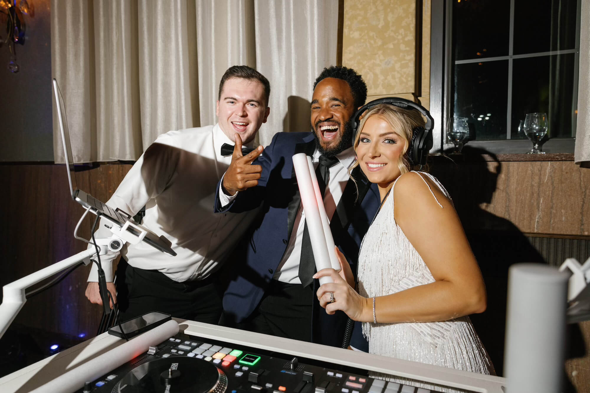 Three people dressed formally stand behind DJ equipment; two men in tuxedos and a woman in a white dress with headphones hold light sticks, smiling and posing for the camera in a well-lit indoor setting.