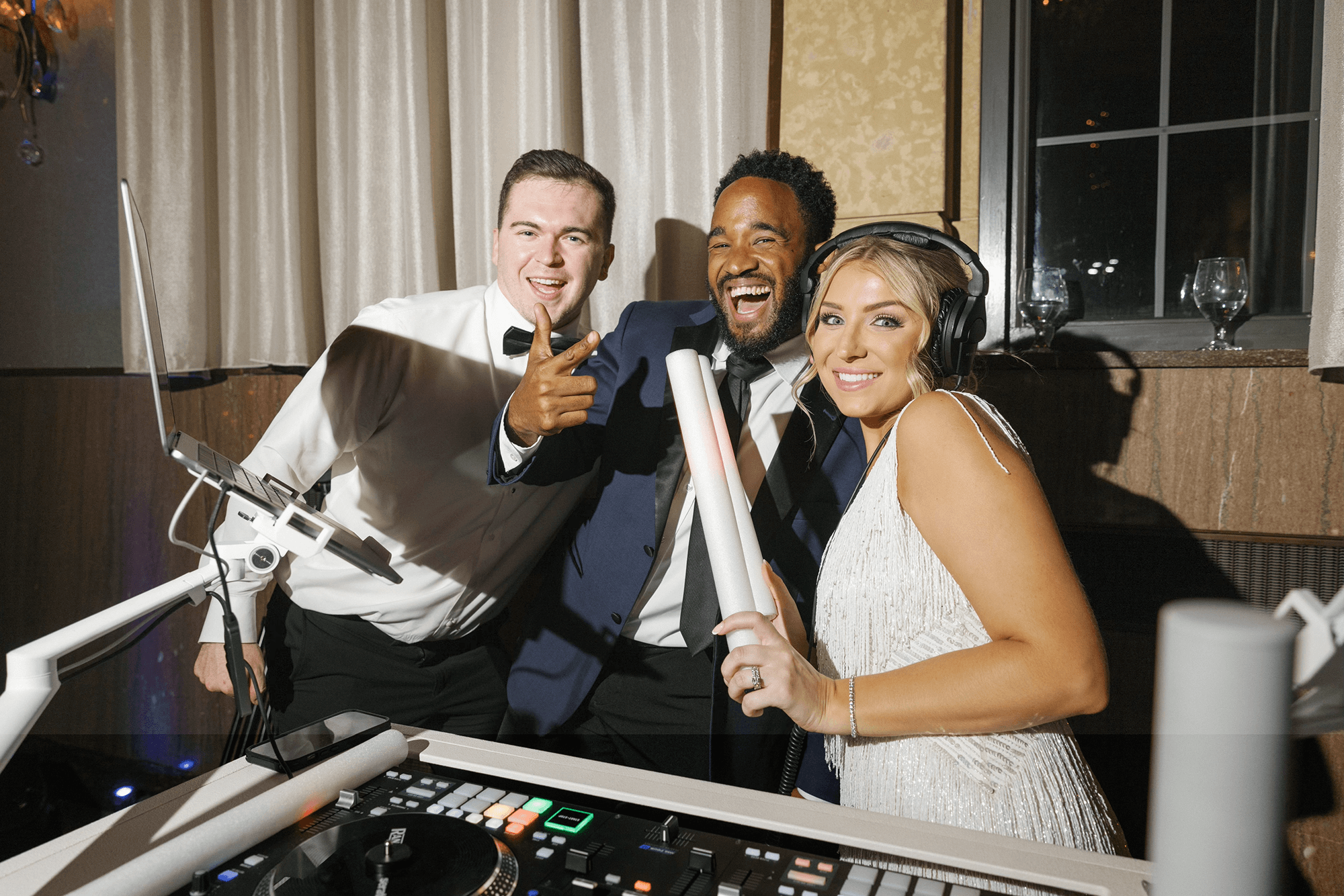 Three people stand behind DJ equipment at an event; one man in a tuxedo, another man in a blue jacket, and a woman in a white dress with headphones, all smiling and posing for the camera.