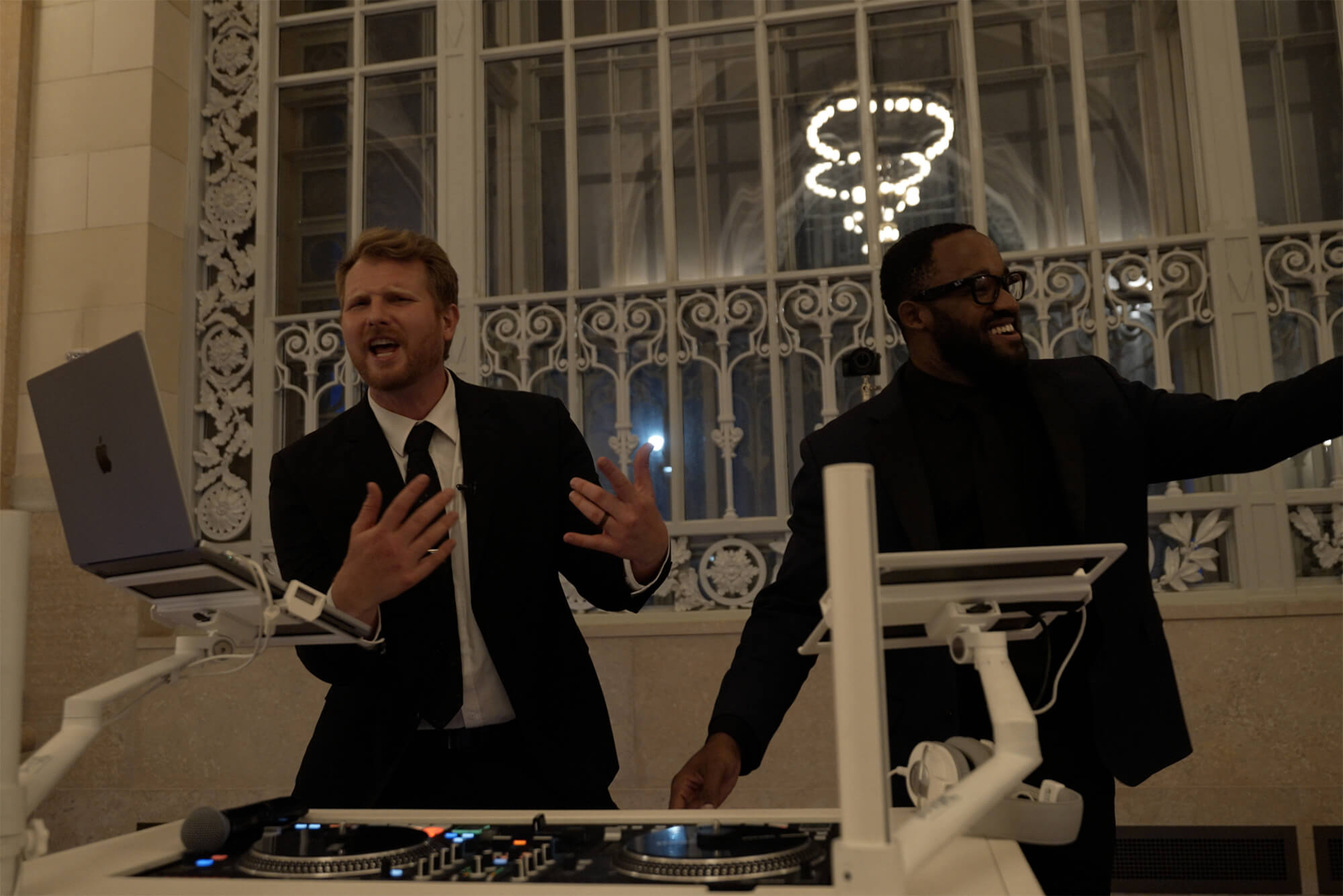 Two men in suits stand behind DJ equipment and a laptop in a well-lit room with large windows and ornate ironwork. One man gestures with his hands while speaking or singing; the other man smiles and raises his arm.