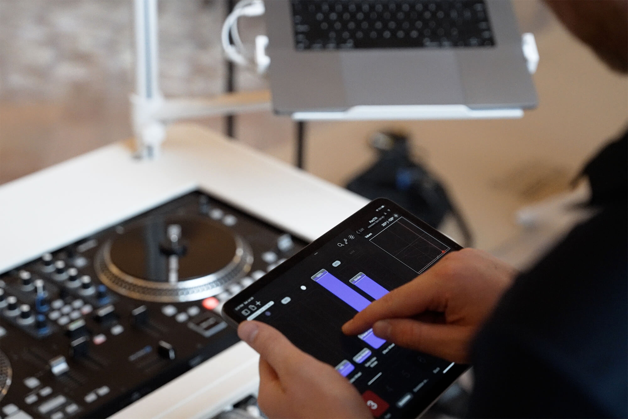 A person uses a tablet with a music app displaying purple bars next to a DJ turntable setup and a laptop on a stand.