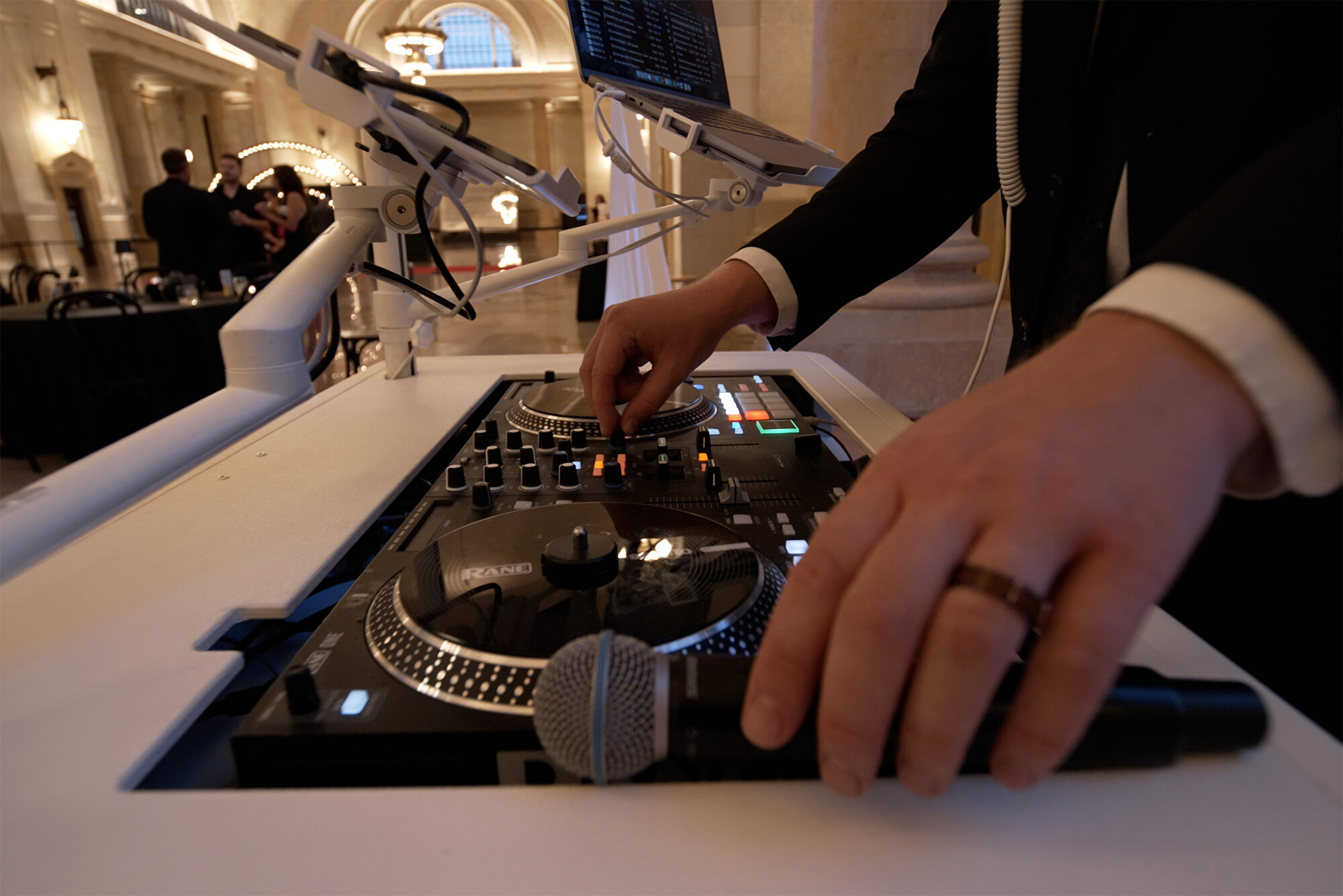 A close-up view of a DJ’s hands adjusting controls on a DJ mixing console with a microphone resting on the surface; people and a chandelier are visible in the blurred background.
