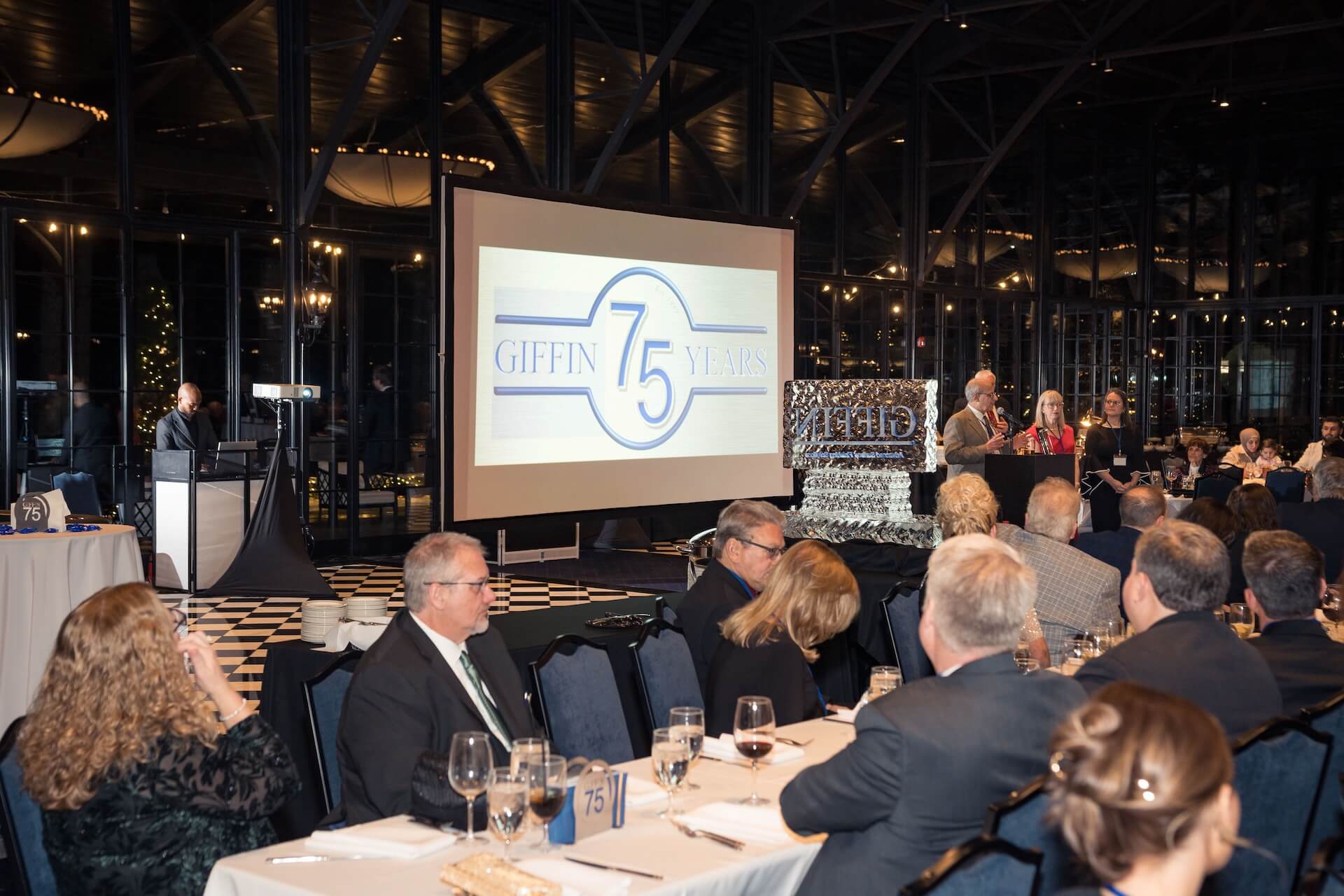 A group of people in formal attire sit at tables during an event. A large screen displays a "Giffin 75 Years" logo. Speakers stand at a podium near a decorative ice sculpture.