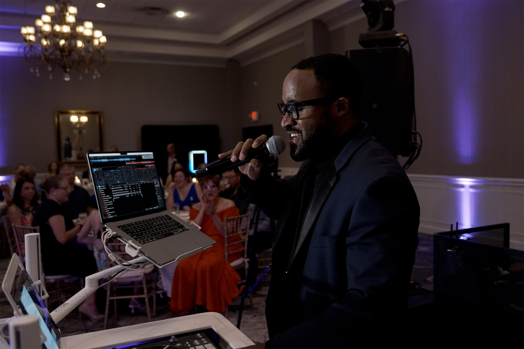 A man wearing glasses and a dark suit holds a microphone and stands behind DJ equipment at an indoor event, with people seated at tables in the background.