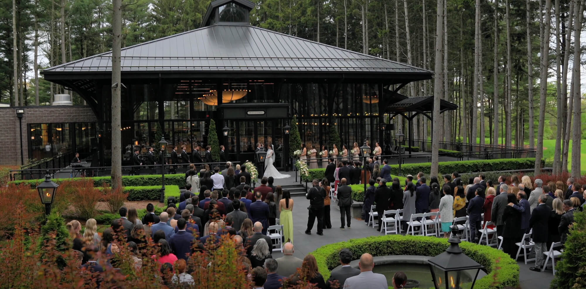 A wedding ceremony is taking place outdoors in front of a glass pavilion surrounded by tall trees, with guests seated on either side of an aisle and the wedding party standing at the front.