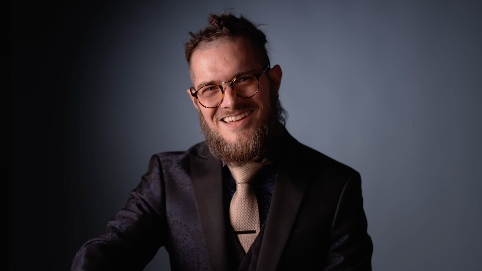 A man with glasses, a beard, and tied-back hair smiles at the camera. He is wearing a dark patterned suit jacket, a light-colored shirt, and a beige tie. The background is a plain, neutral color.