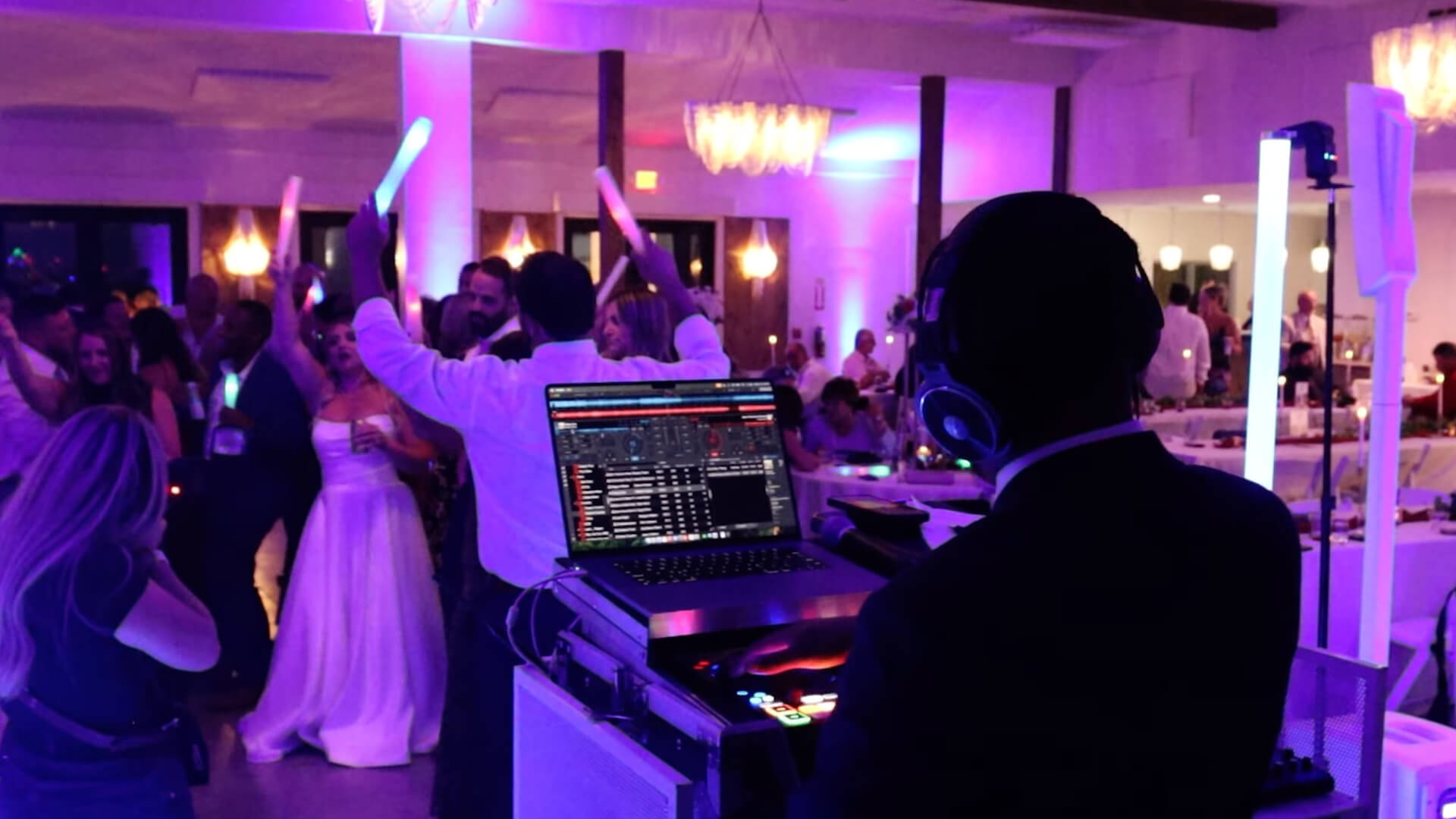 A DJ wearing headphones stands at a booth with a laptop, playing music for a group of people dancing at an indoor event with purple lighting. A bride is visible on the dance floor among the guests.