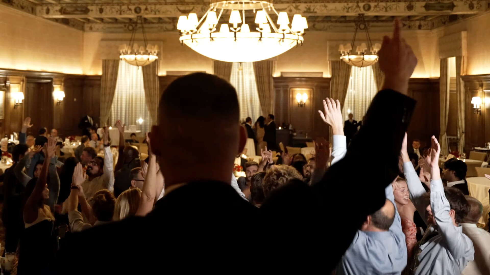 A person in a suit stands with an arm raised, facing a large crowd with raised hands in an elegant, chandelier-lit banquet hall. The focus is on the back of the person, with the audience and room in view.
