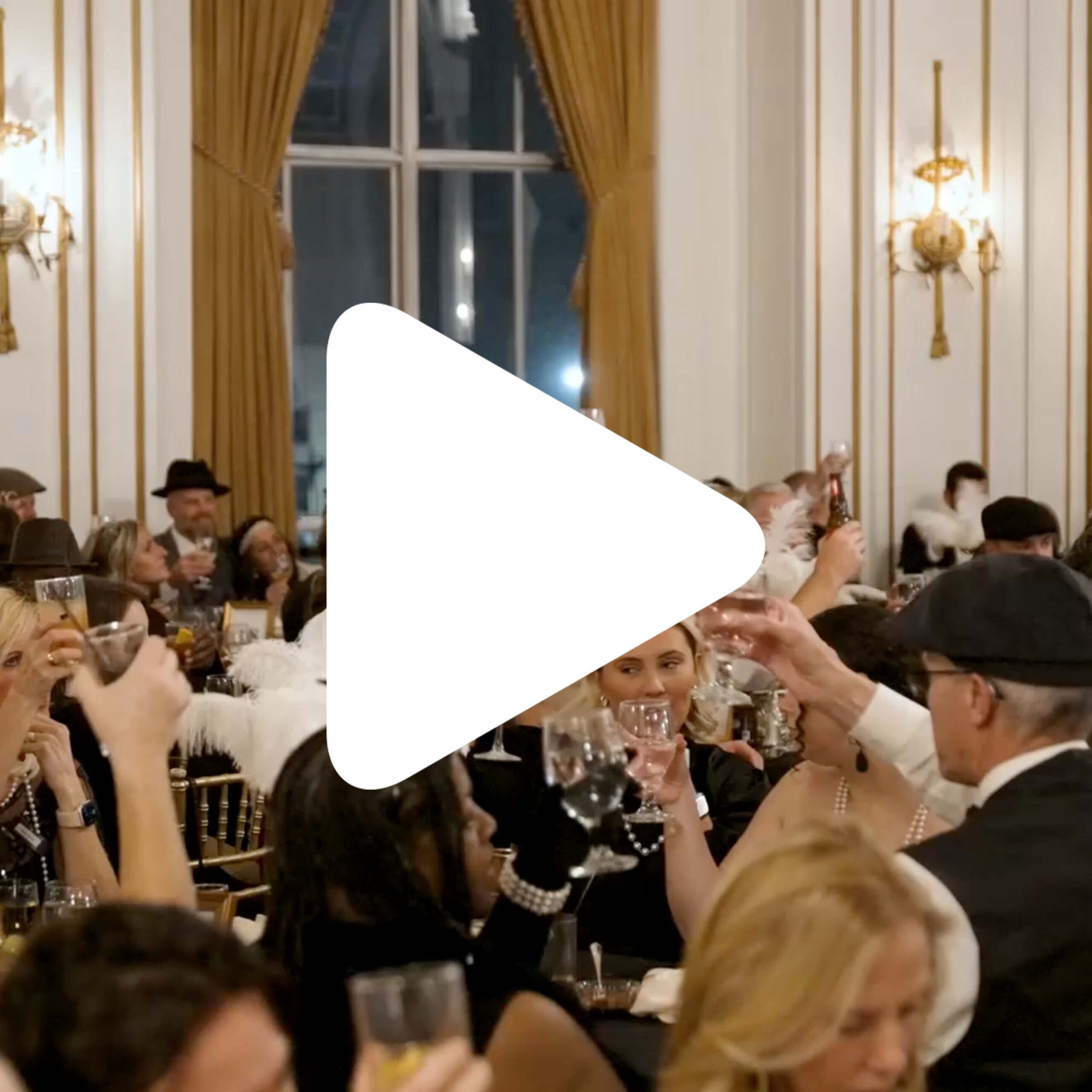 A large group of people dressed in formal attire raise glasses in a toast at a banquet hall with gold accents and tall curtains. A large play button icon is centered over the image.