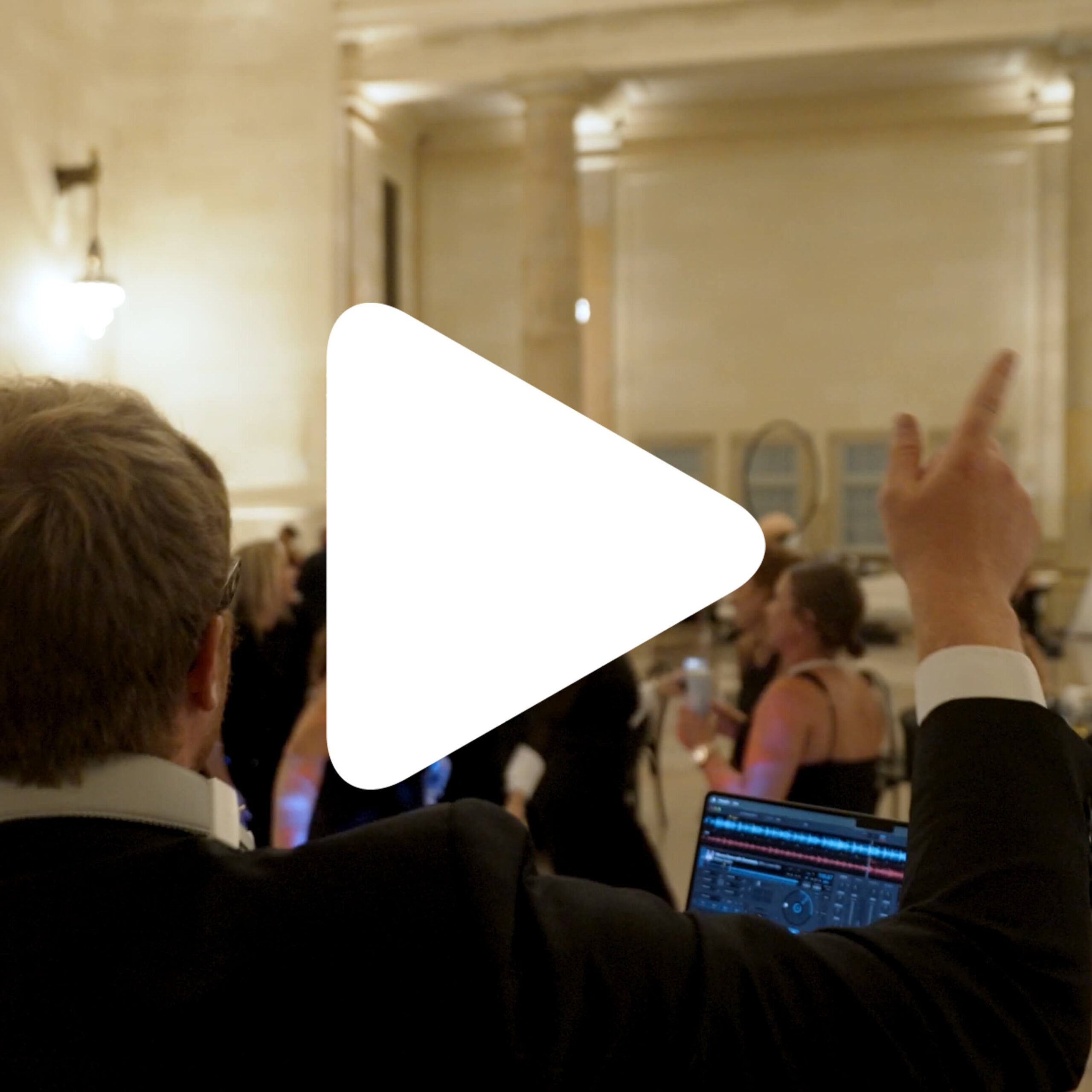 A person in formal attire stands near DJ equipment, raising one hand, while several people dance in the background inside a large, well-lit hall. A large play button icon is centered over the image.