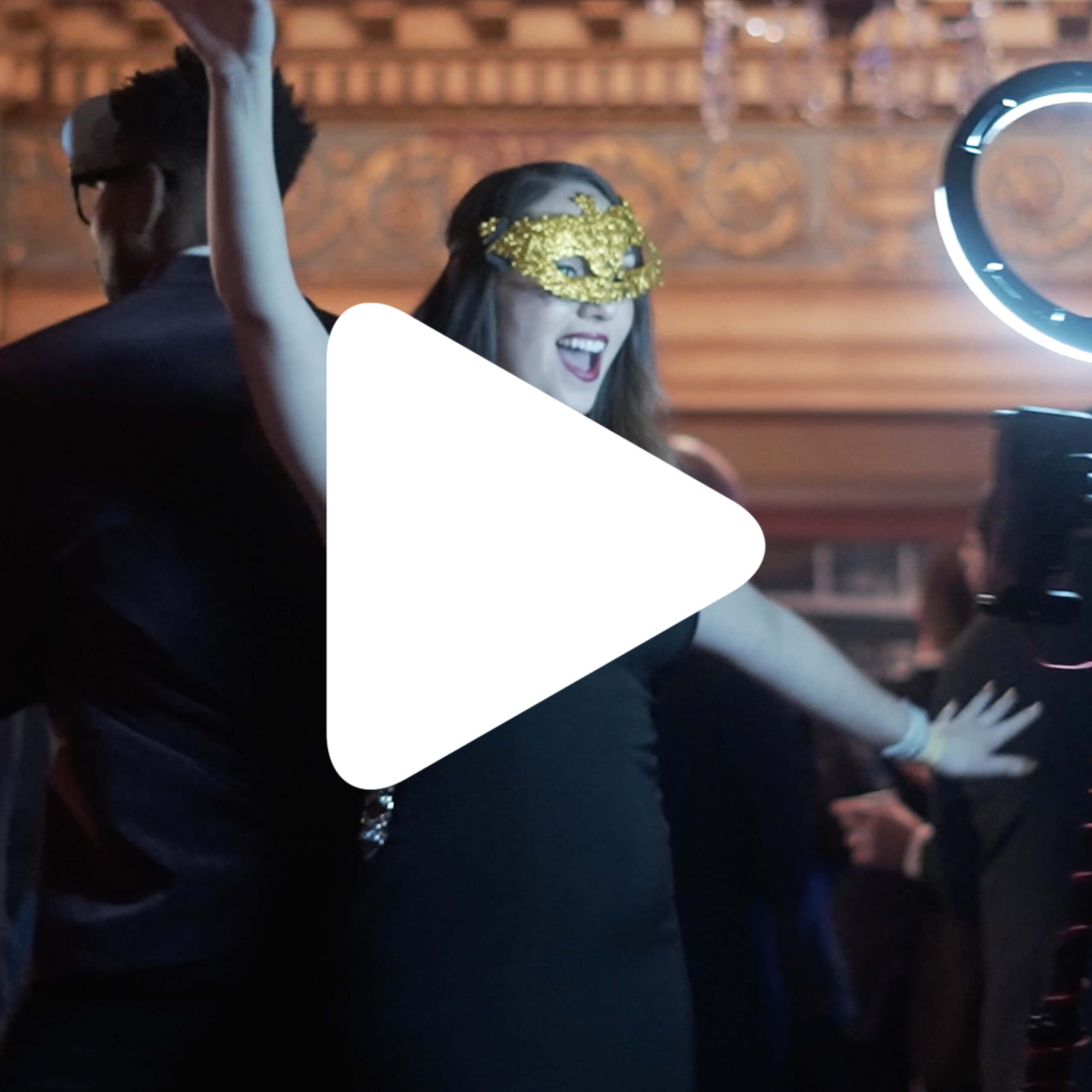 A person wearing a gold masquerade mask and black outfit stands with arms raised at a lively indoor event. Other people and a ring light are visible in the background. A large play button icon overlays the image.