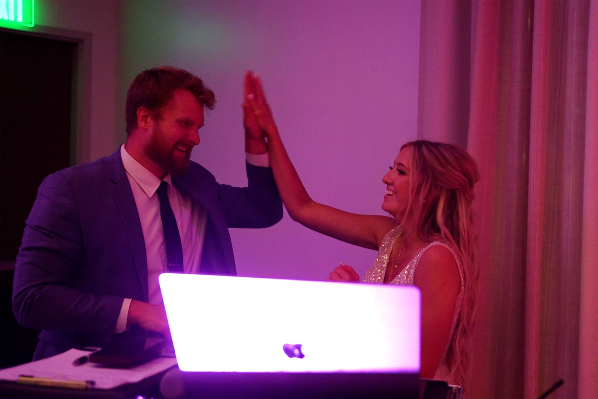 A man in a suit and a woman in a sleeveless dress high-five each other in a dimly lit room with purple lighting. A glowing laptop is on the table in front of them.