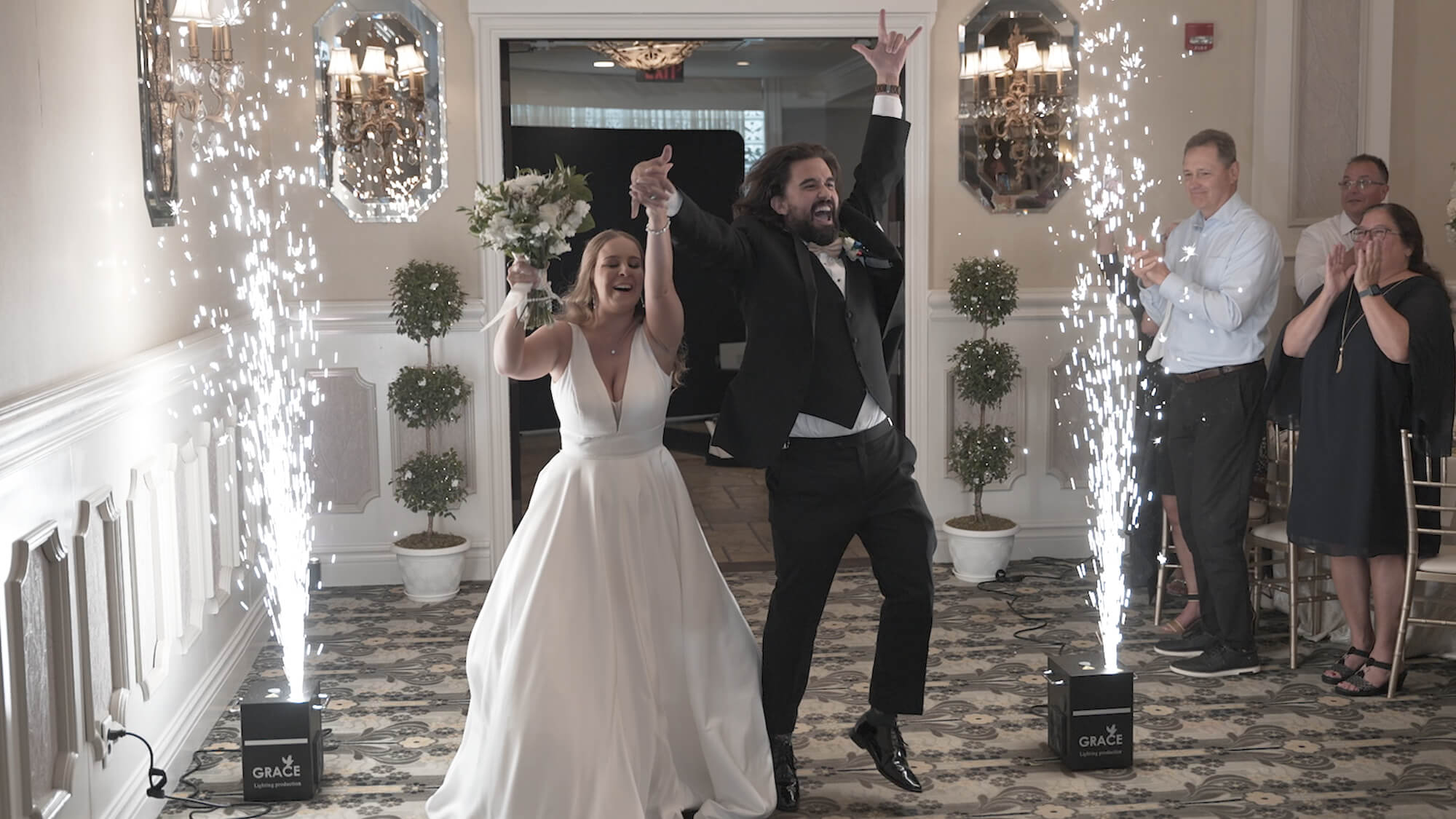 A newlywed couple enters a room, holding hands and celebrating, with indoor sparklers on either side. The bride wears a white gown and holds a bouquet; the groom wears a suit. Guests stand and applaud in the background.