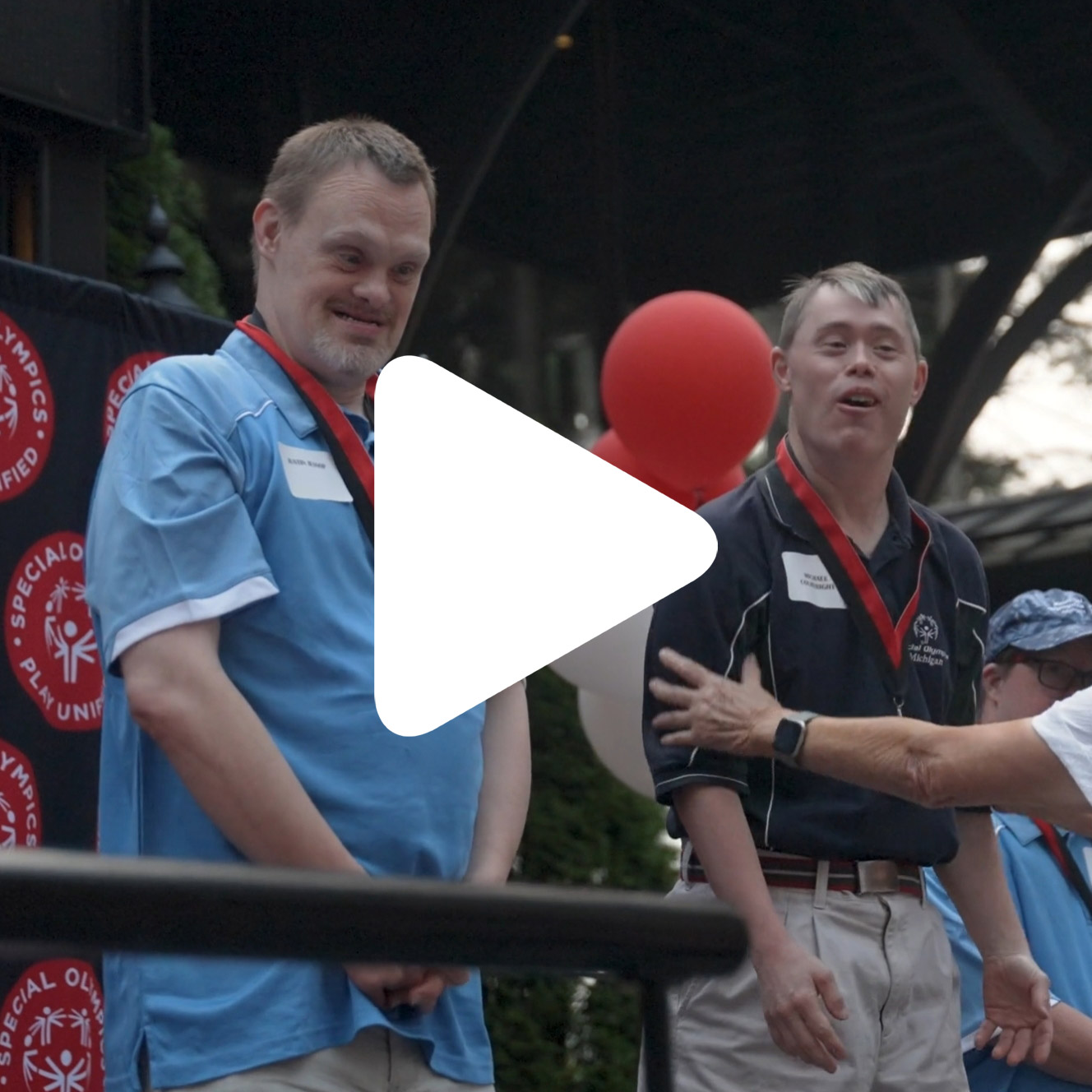 Two men wearing medals stand on a stage at a Special Olympics event, with one person reaching toward them. There are red and white balloons and a banner in the background. A white play button icon is overlaid on the image.