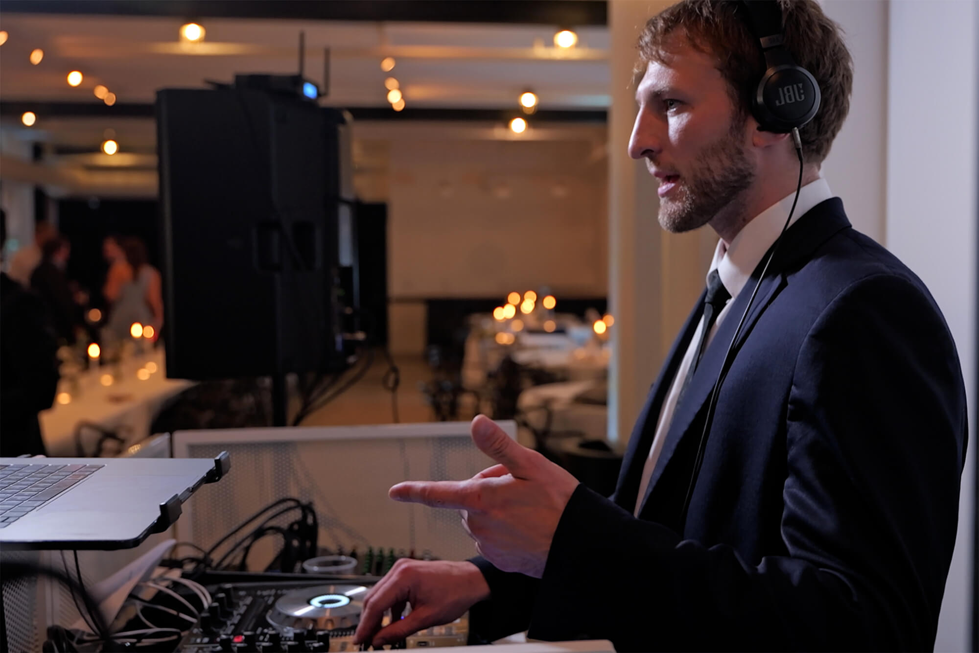 A man in a suit and headphones stands at a DJ booth, operating a mixer and pointing with his right hand. In the background, tables with candles and people in formal attire are visible.
