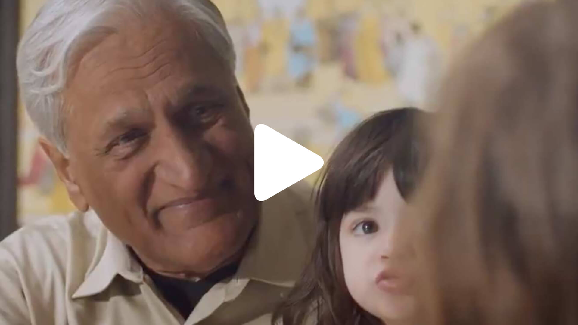 An older man with gray hair and a light shirt smiles while sitting beside a young child with long dark hair. Part of another person’s head is visible in the foreground. A blurred background shows people and colors.