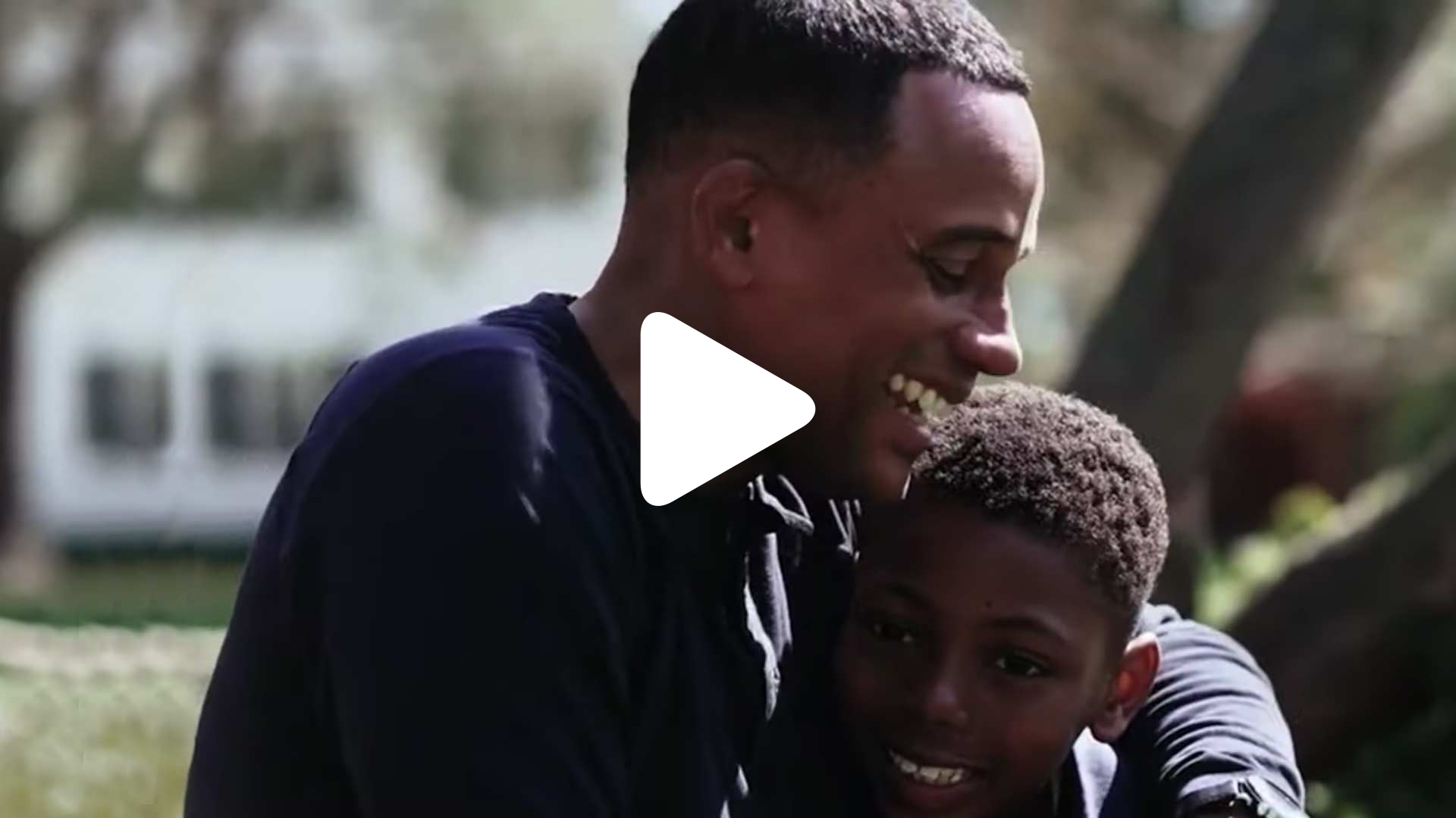 A man smiles and hugs a young boy outdoors. Both appear happy and are dressed in dark clothing. Sunlight and greenery are visible in the background. A white play button is centered on the image.
